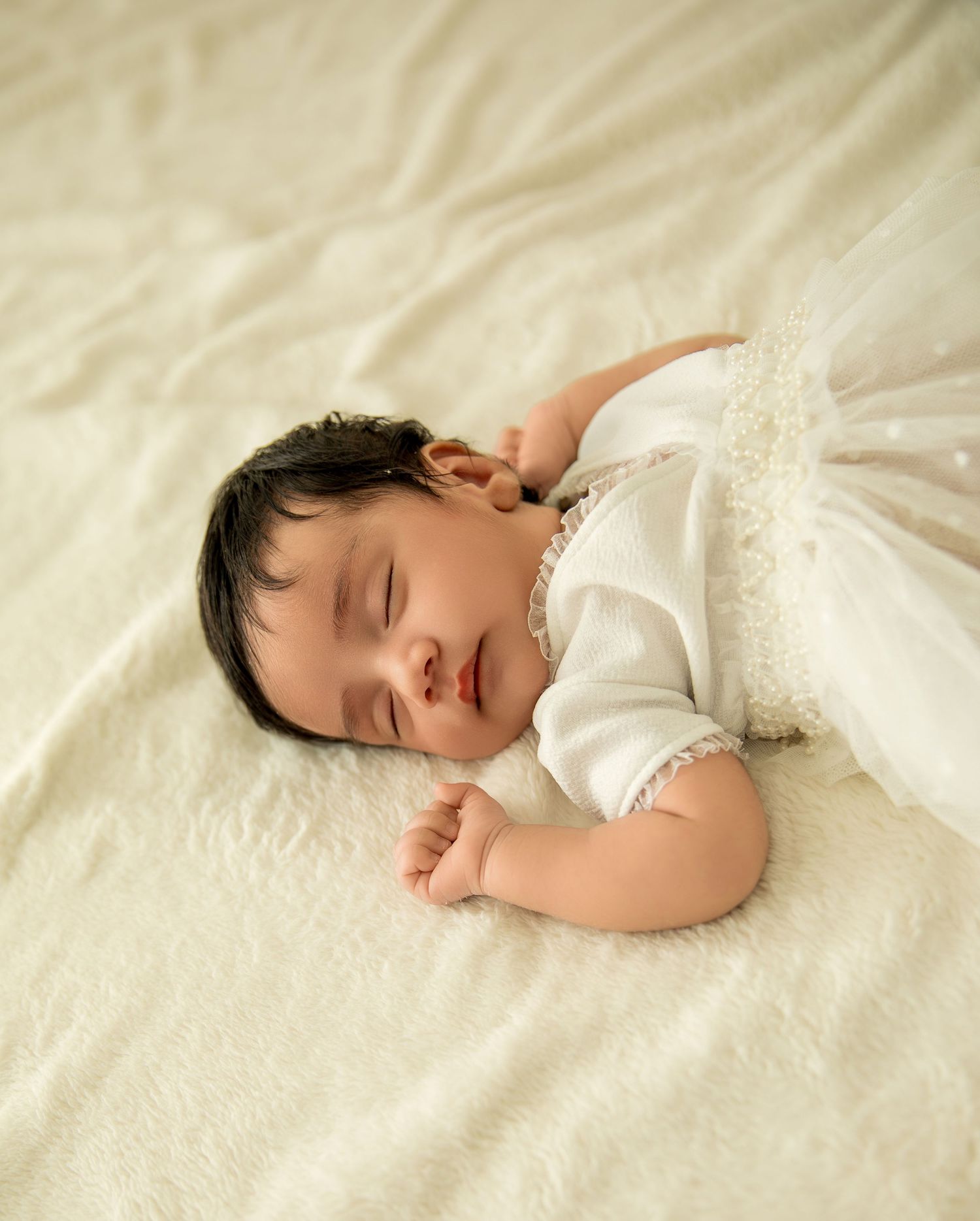A sleeping baby in white lace clothing rests peacefully on cream-colored bedding.