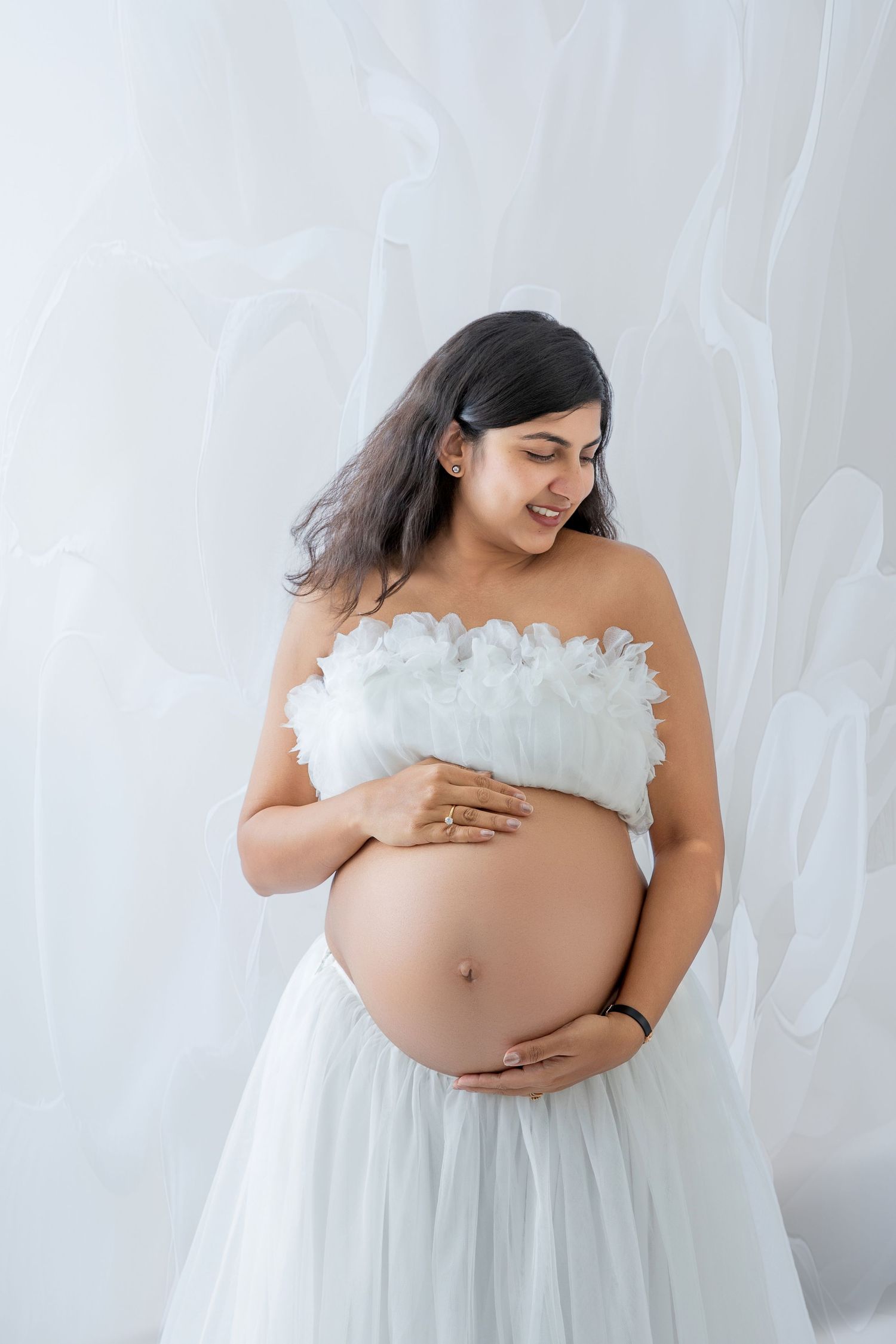 Smiling pregnant woman in white feathered tube dress cradling her baby bump against a soft white background.