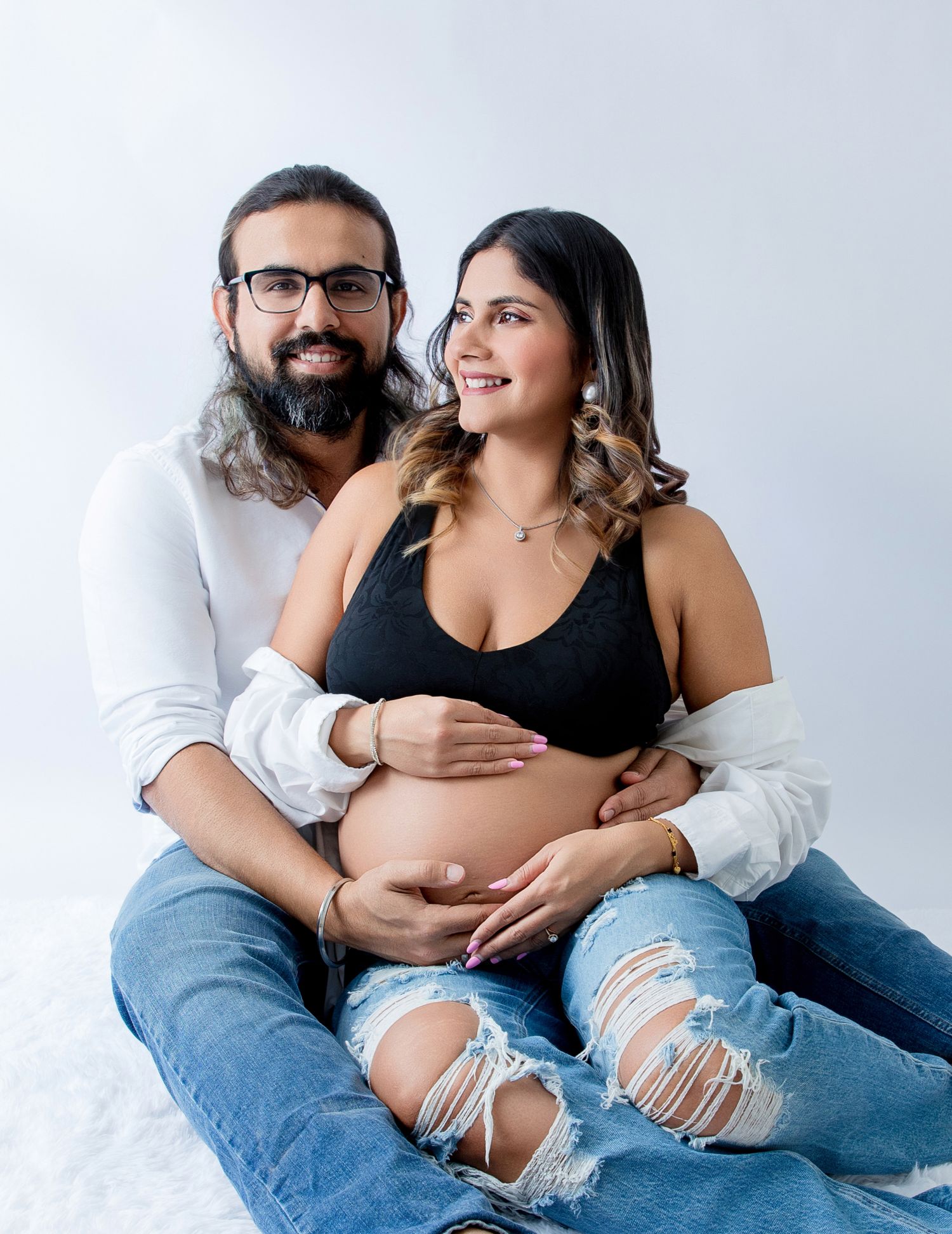 Couple posing for maternity photo shoot, pregnant woman in black crop top and ripped jeans, man with beard and glasses.