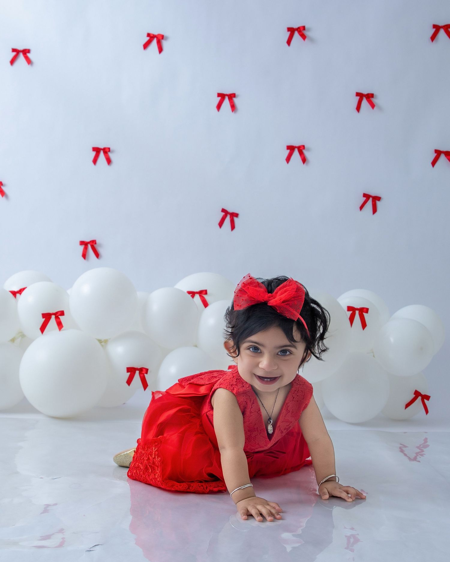 Baby in red dress lying on floor with white balloons and red bows in background.