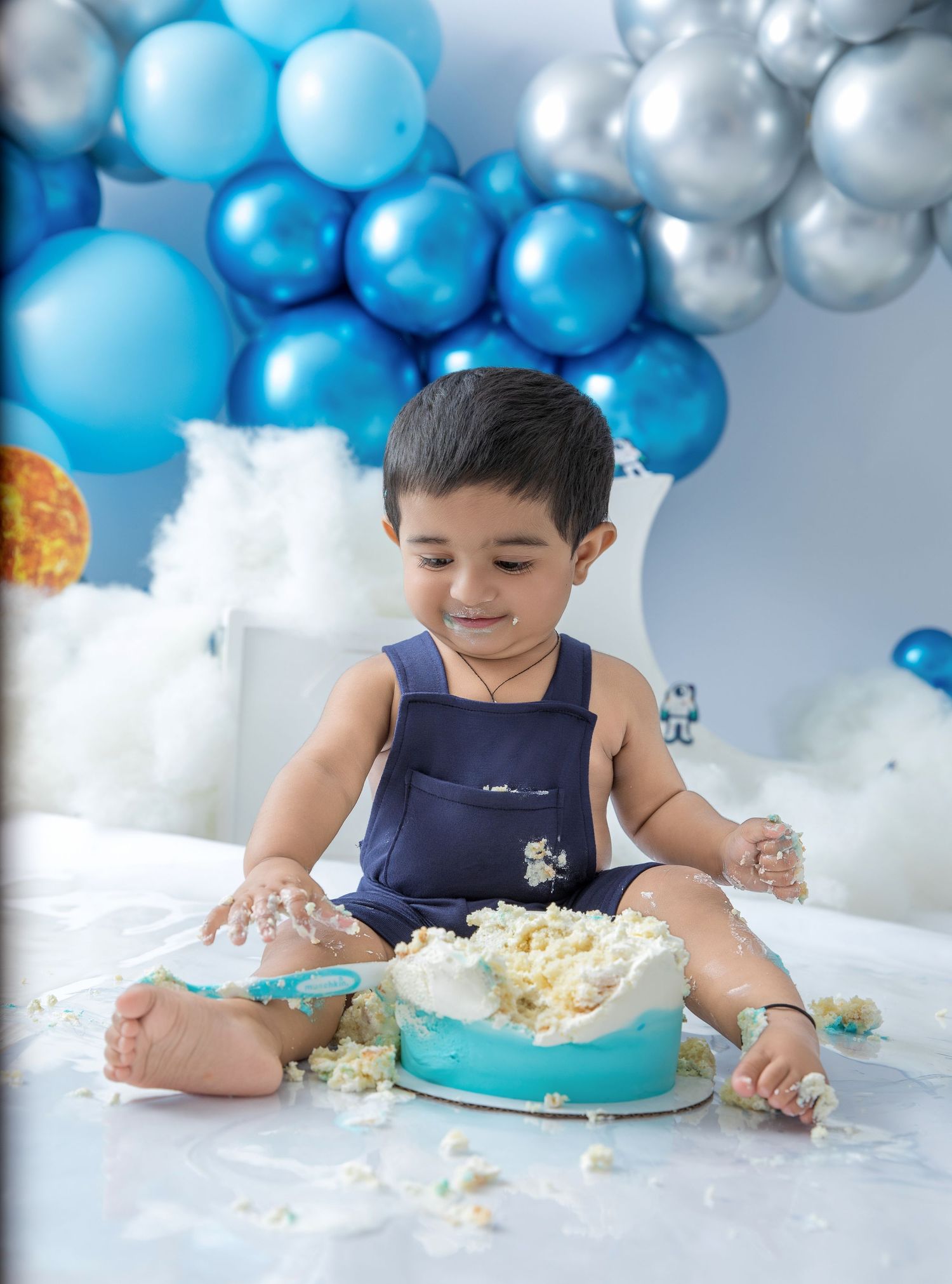Baby in navy outfit enjoying a cake smash photo session with blue and silver balloon decorations.