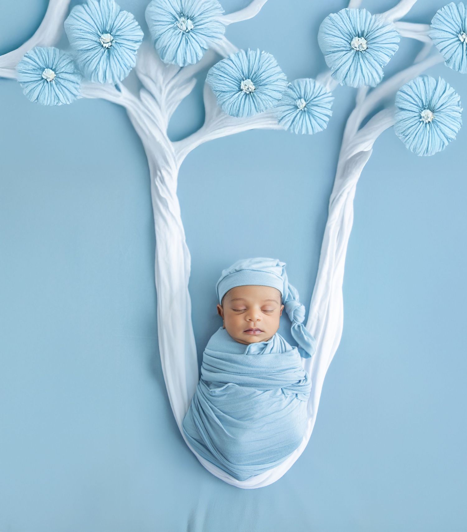 Creative newborn photography with white paper trees and blue flowers on light blue background.