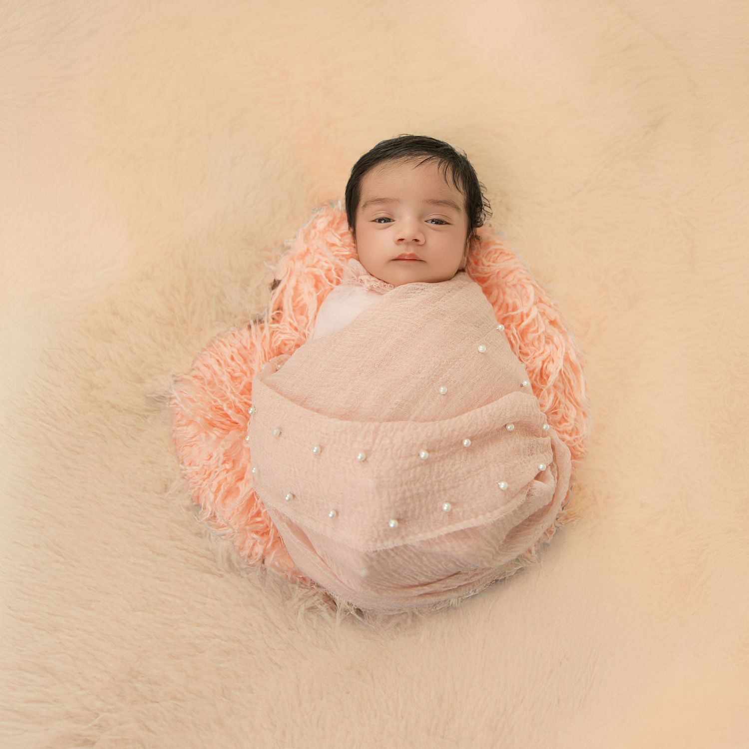 Sleeping newborn baby in a peach-colored wrap photographed from above on a cream-colored background.