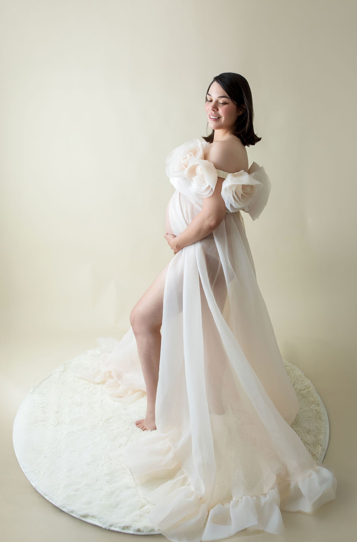 Pregnant woman in sheer white off-shoulder gown with ruffled sleeves poses gracefully in a studio maternity photo.