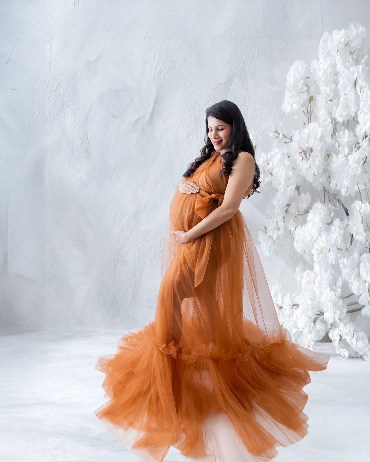 Pregnant woman in elegant rust orange tulle maternity gown posing gracefully in a white studio with floral backdrop.