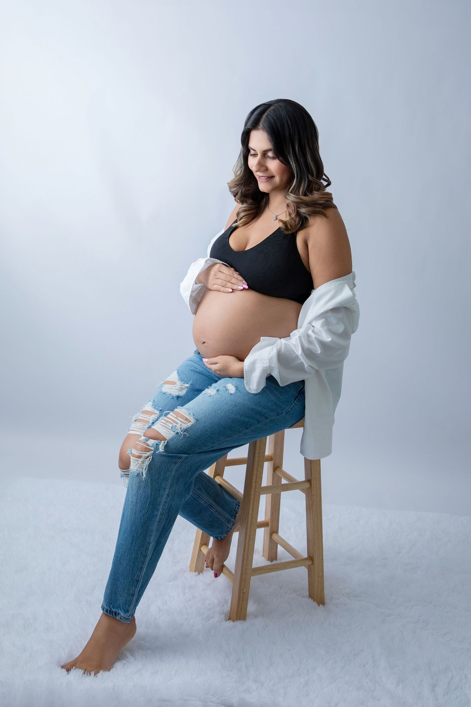 Pregnant woman in black crop top and ripped jeans sitting on wooden stool, cradling baby bump in studio setting.