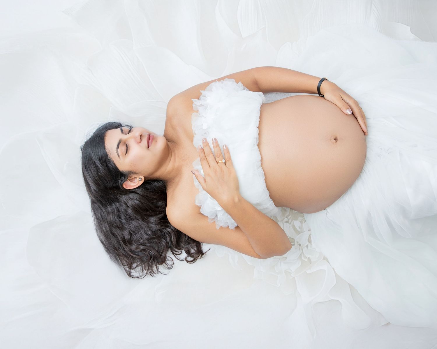 Pregnant woman in white feathered dress lying on white fabric, cradling baby bump during maternity photo shoot.