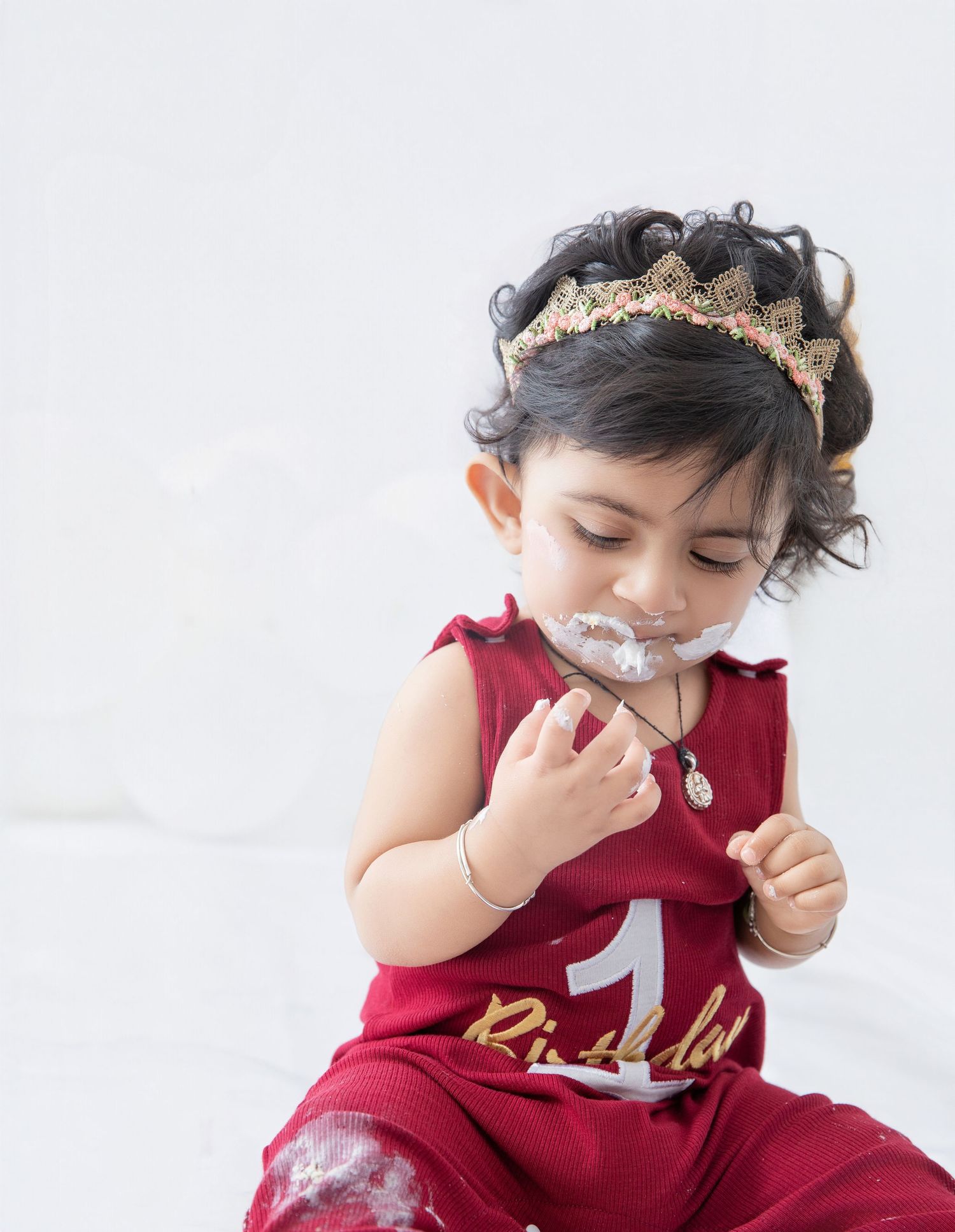 Baby in burgundy outfit with gold crown tastes cake frosting during celebration shoot.