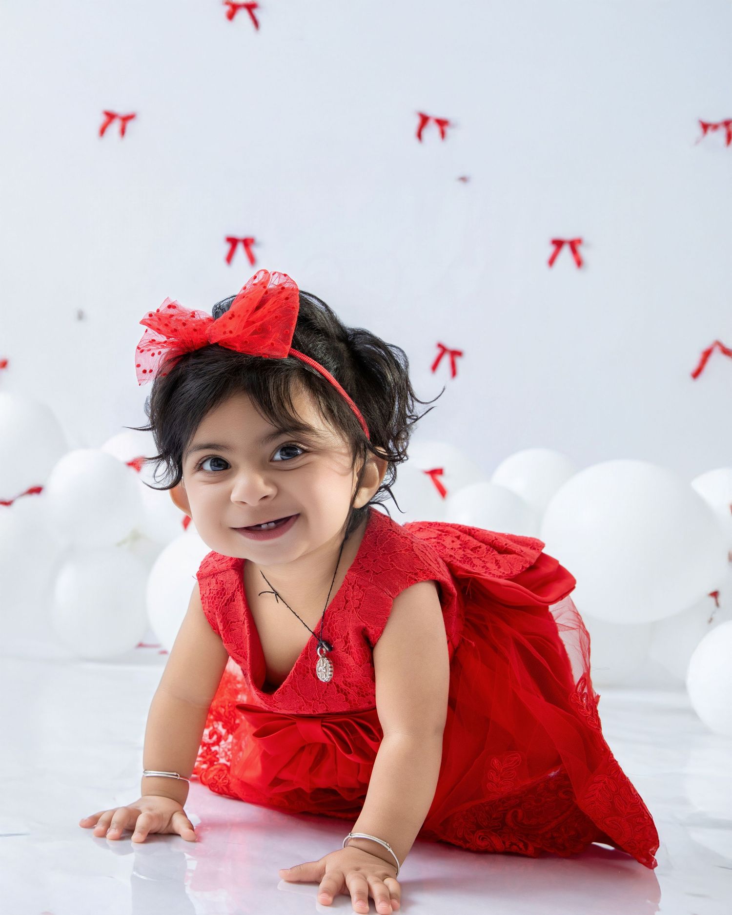 Baby in red dress crawls and plays on white floor with red bow decorations in background.