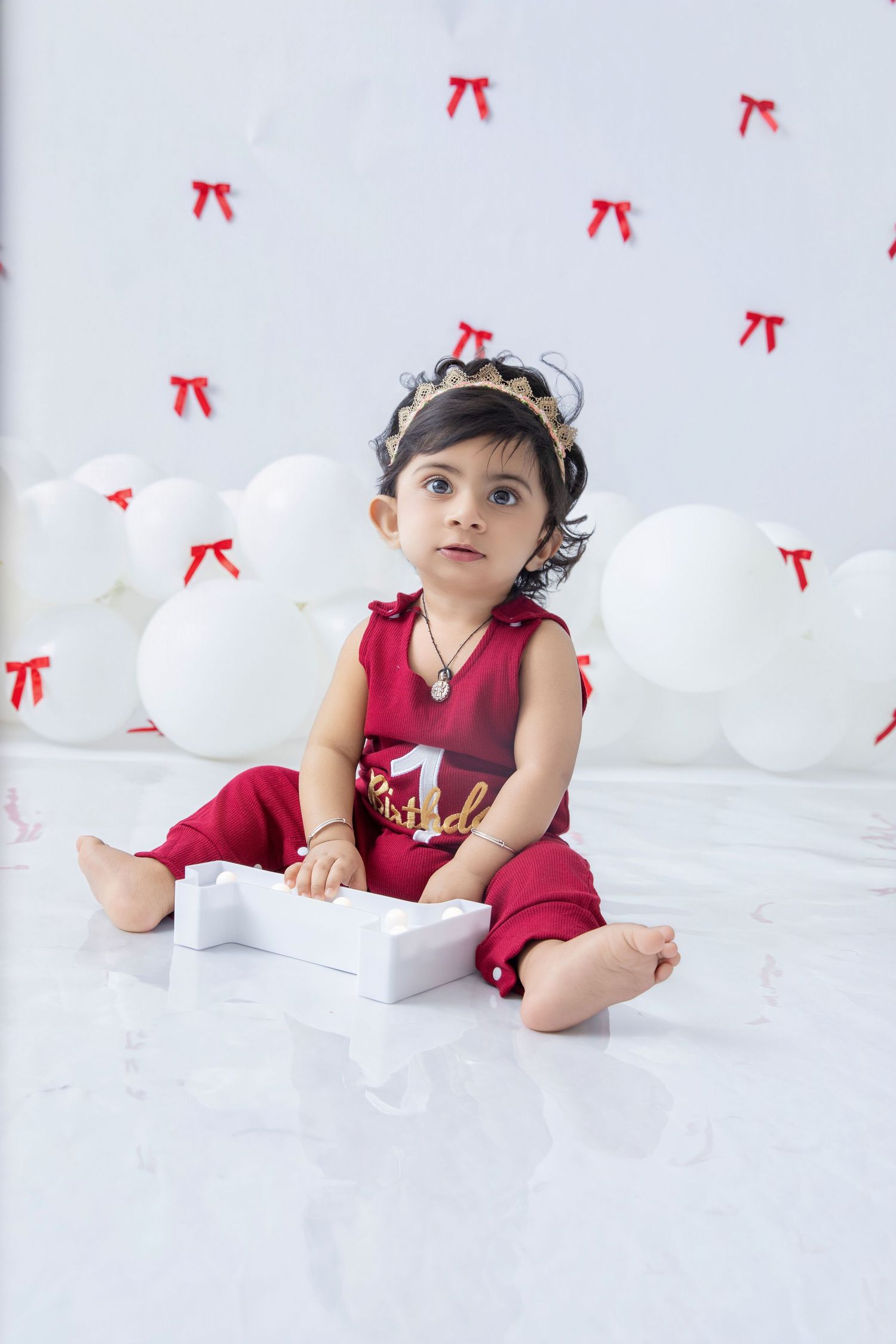 Baby in red outfit lies on white surface with decorative bows in background.