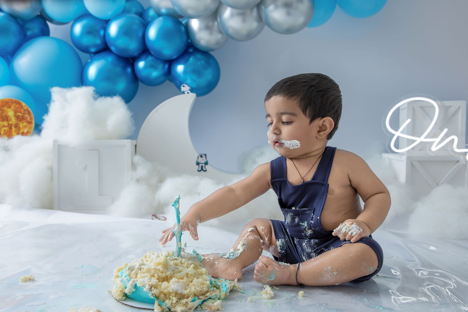Baby plays with cake on white floor surrounded by blue and silver balloons during cake smash session.