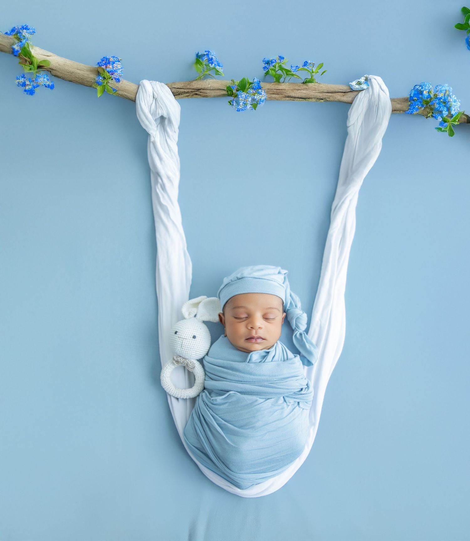 Newborn baby swaddled in light blue fabric arranged in creative letter shapes against a blue background with tiny flowers.