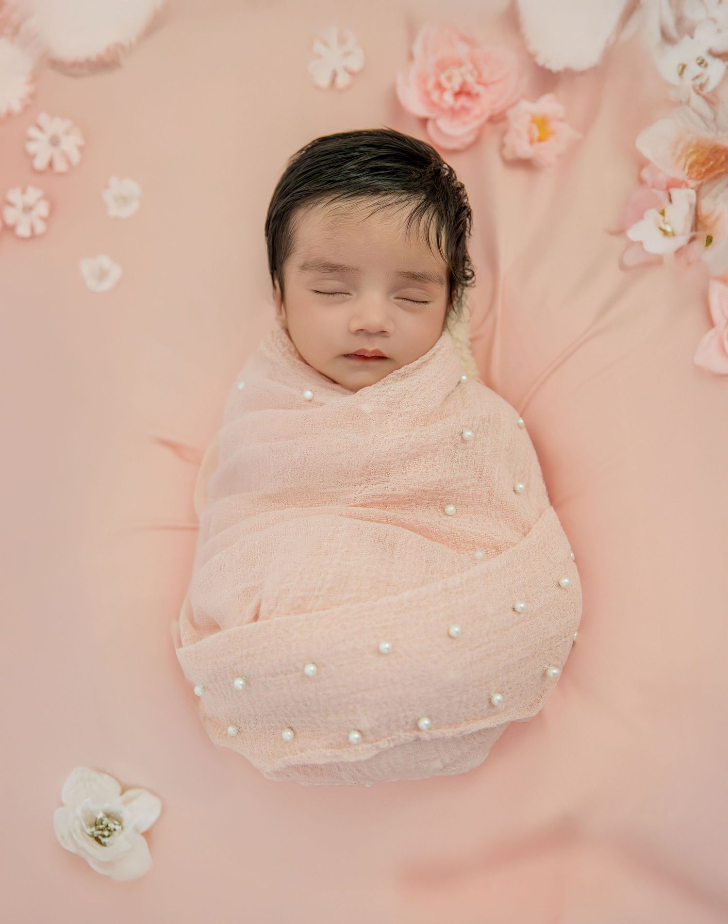 Sleeping newborn baby wrapped in soft pink blanket with delicate floral decorations and pearls in dreamy nursery setting.