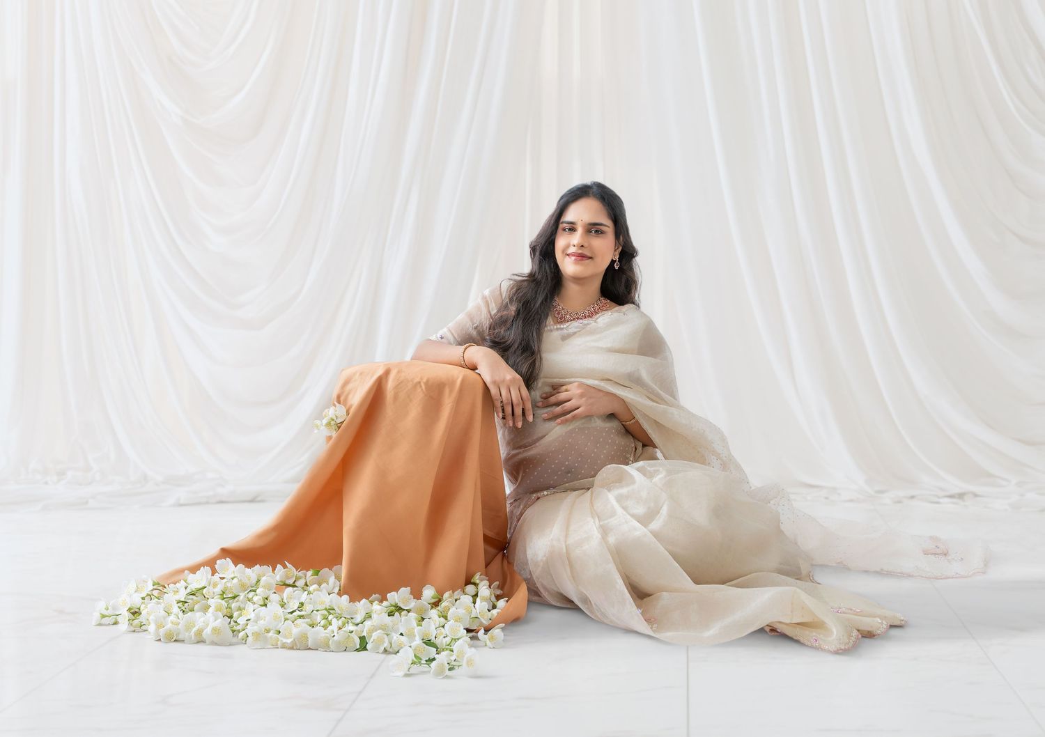 Pregnant woman in elegant cream and orange saree sitting gracefully against white draped backdrop with white flowers.