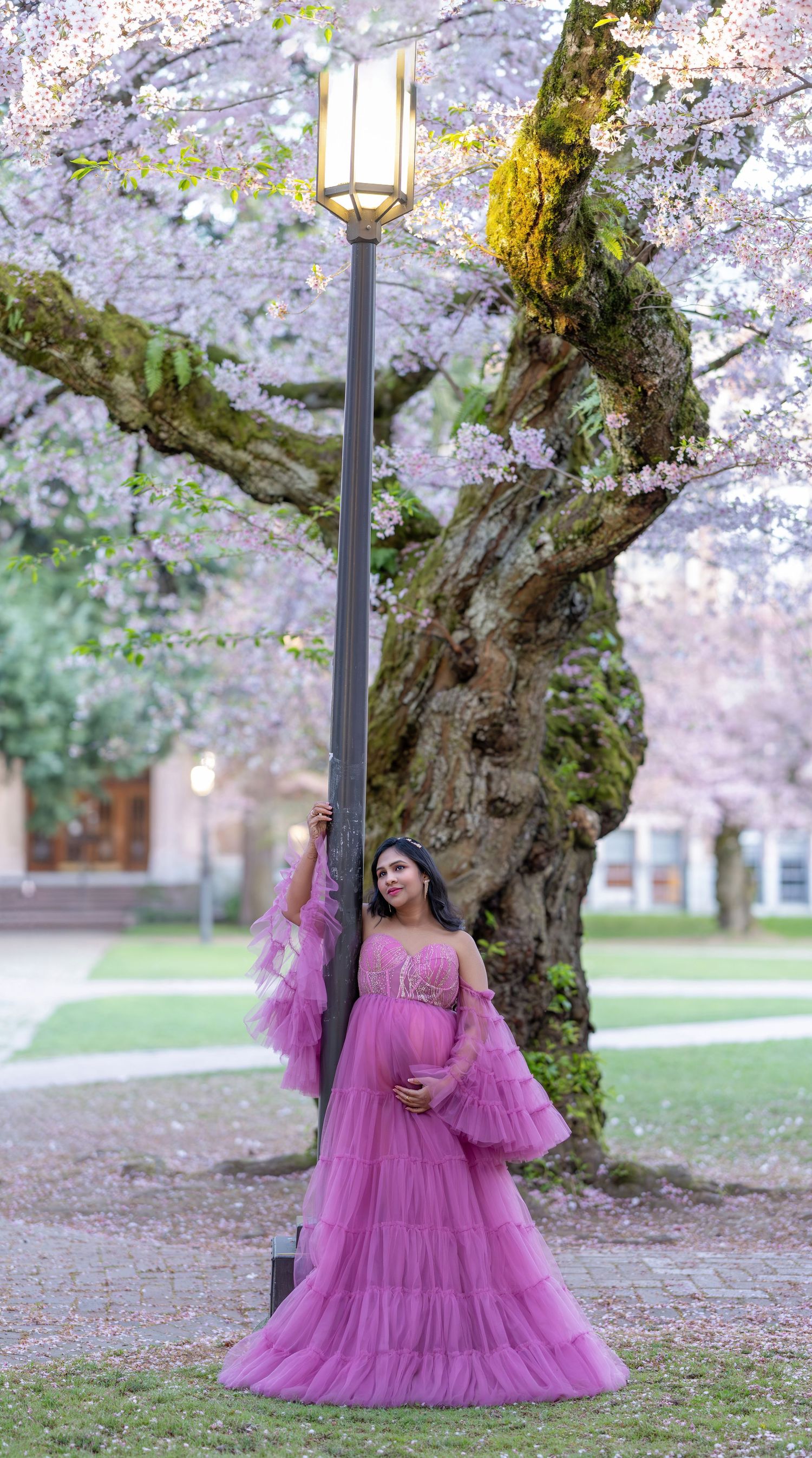 Pregnant woman in pink tulle gown poses by lamppost under blooming cherry blossom trees.