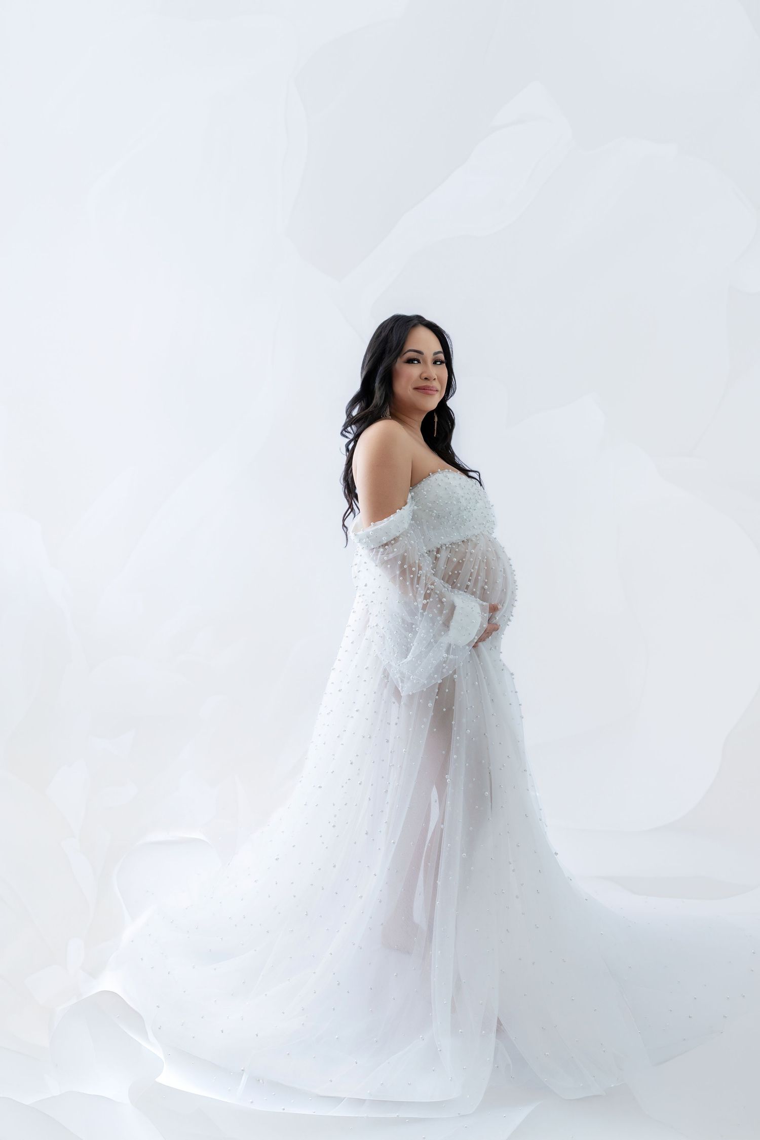 Pregnant woman in flowing white tulle maternity gown posing gracefully against a soft white studio background.