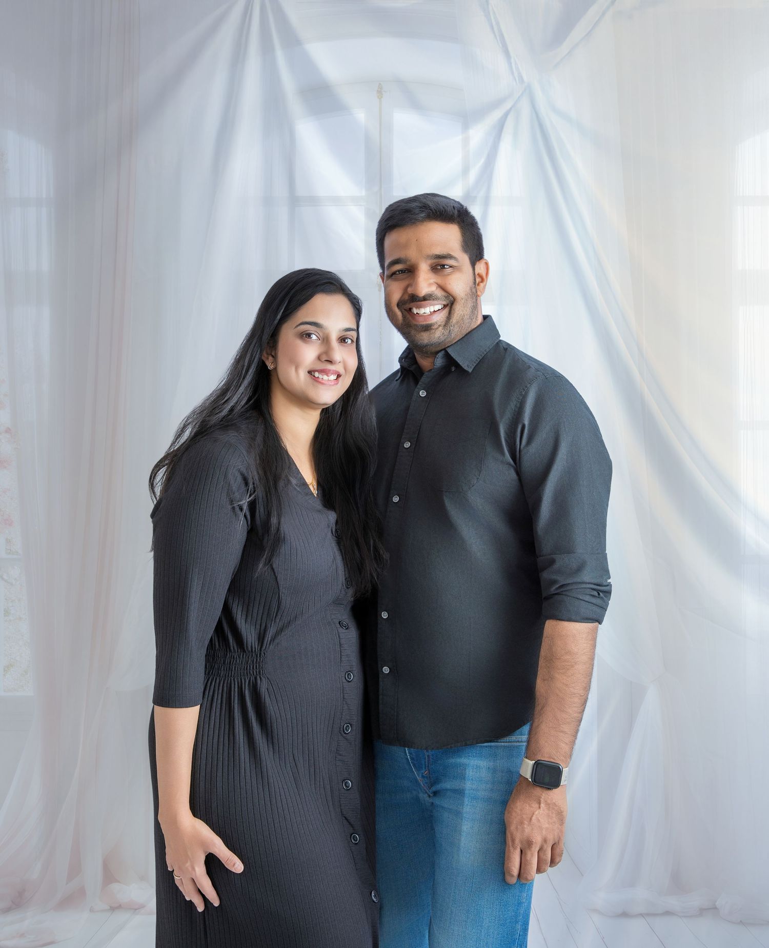 A couple in coordinating black outfits poses together against a white backdrop in a series of casual studio photos.