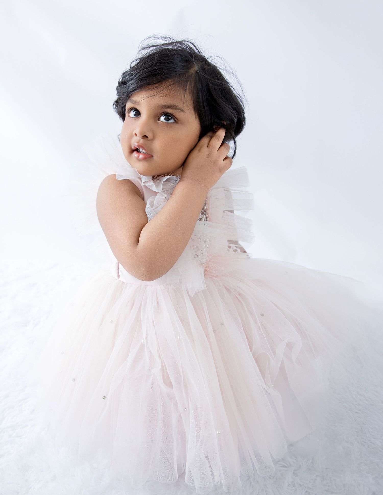 Young child in delicate white tulle dress touching their hair during photo session.