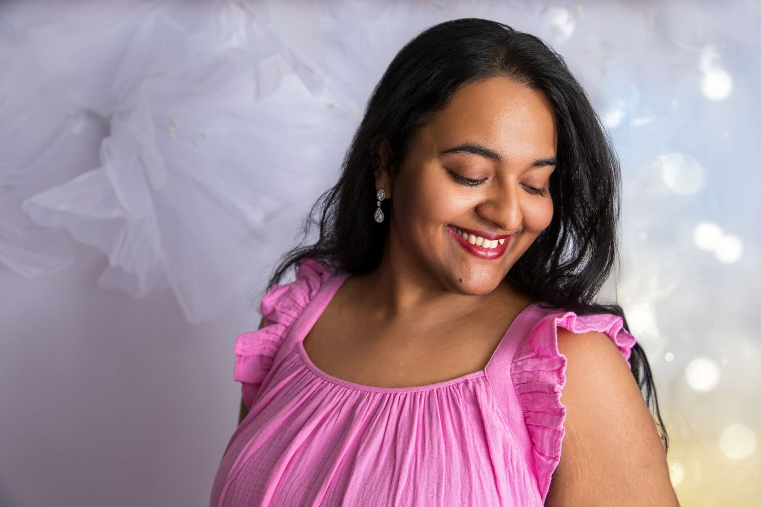 Woman wearing pink ruffled top poses against light background with bokeh effects.