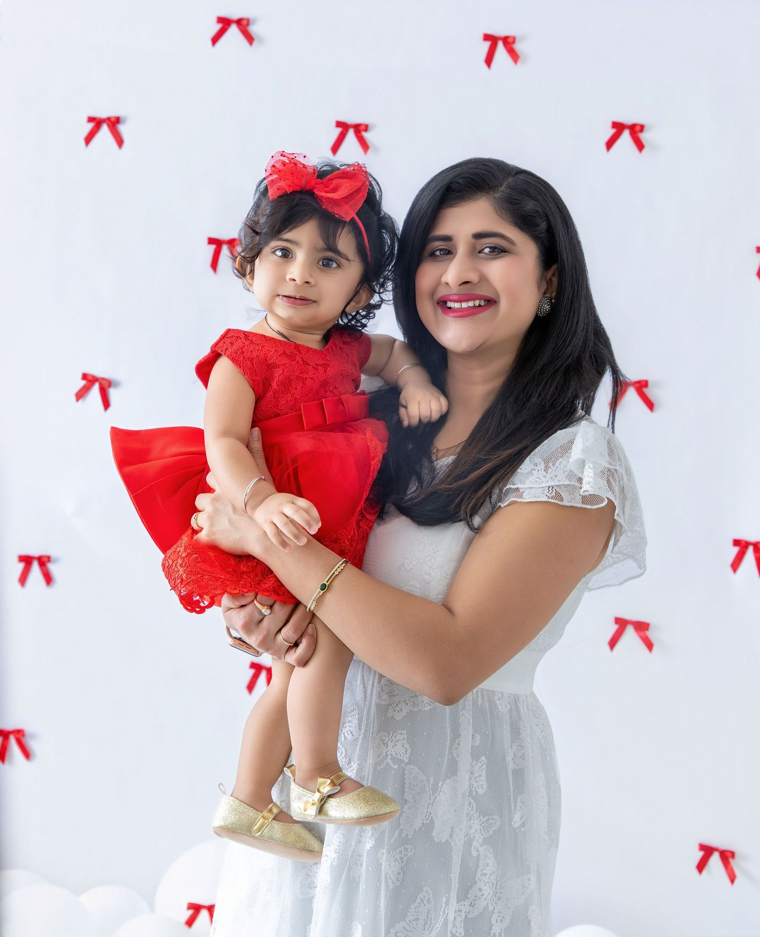 A toddler in a red dress with matching bow poses with adult against a white background with red bow pattern.