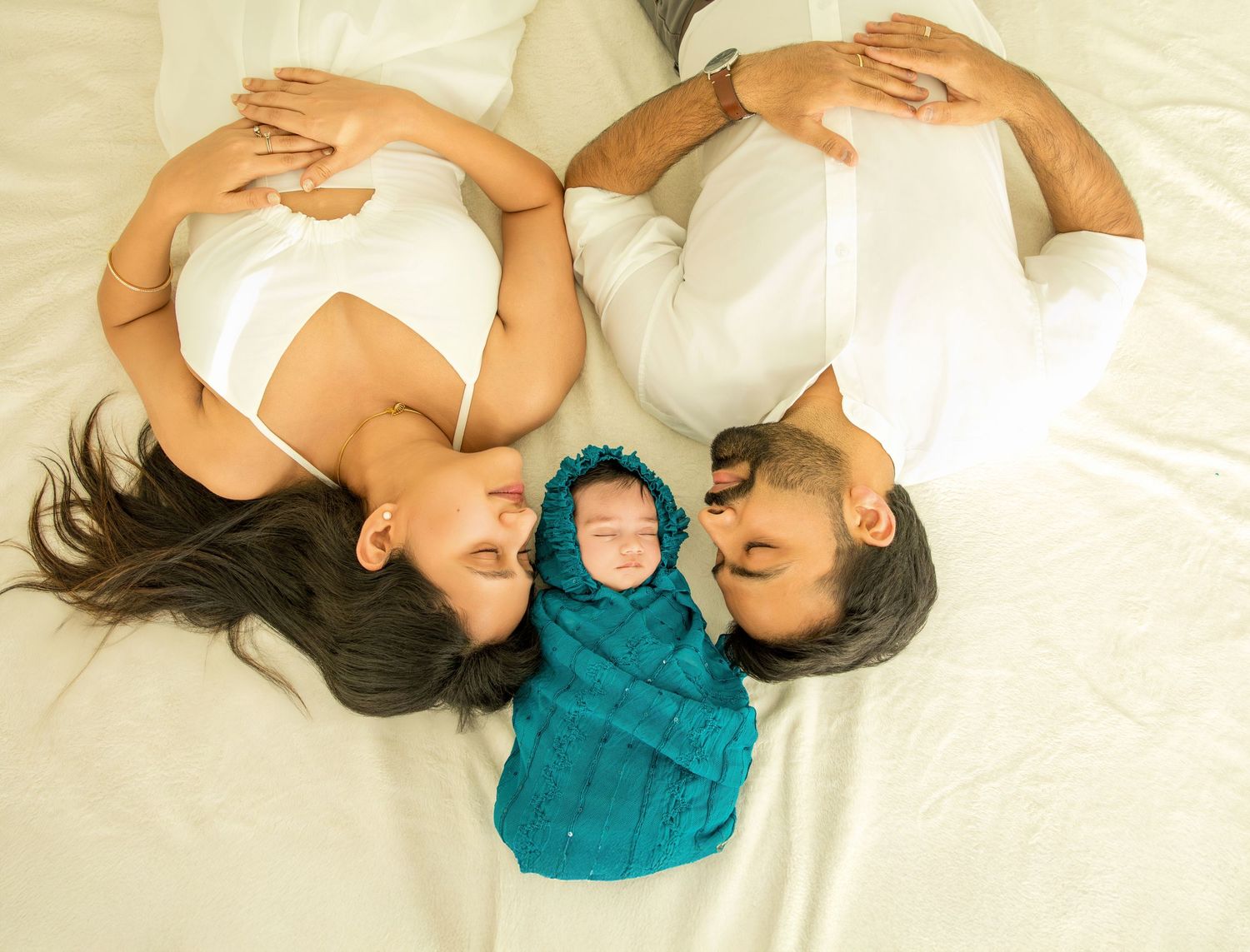 Family lying on white bedding with newborn baby in teal swaddle creating heart shape formation.