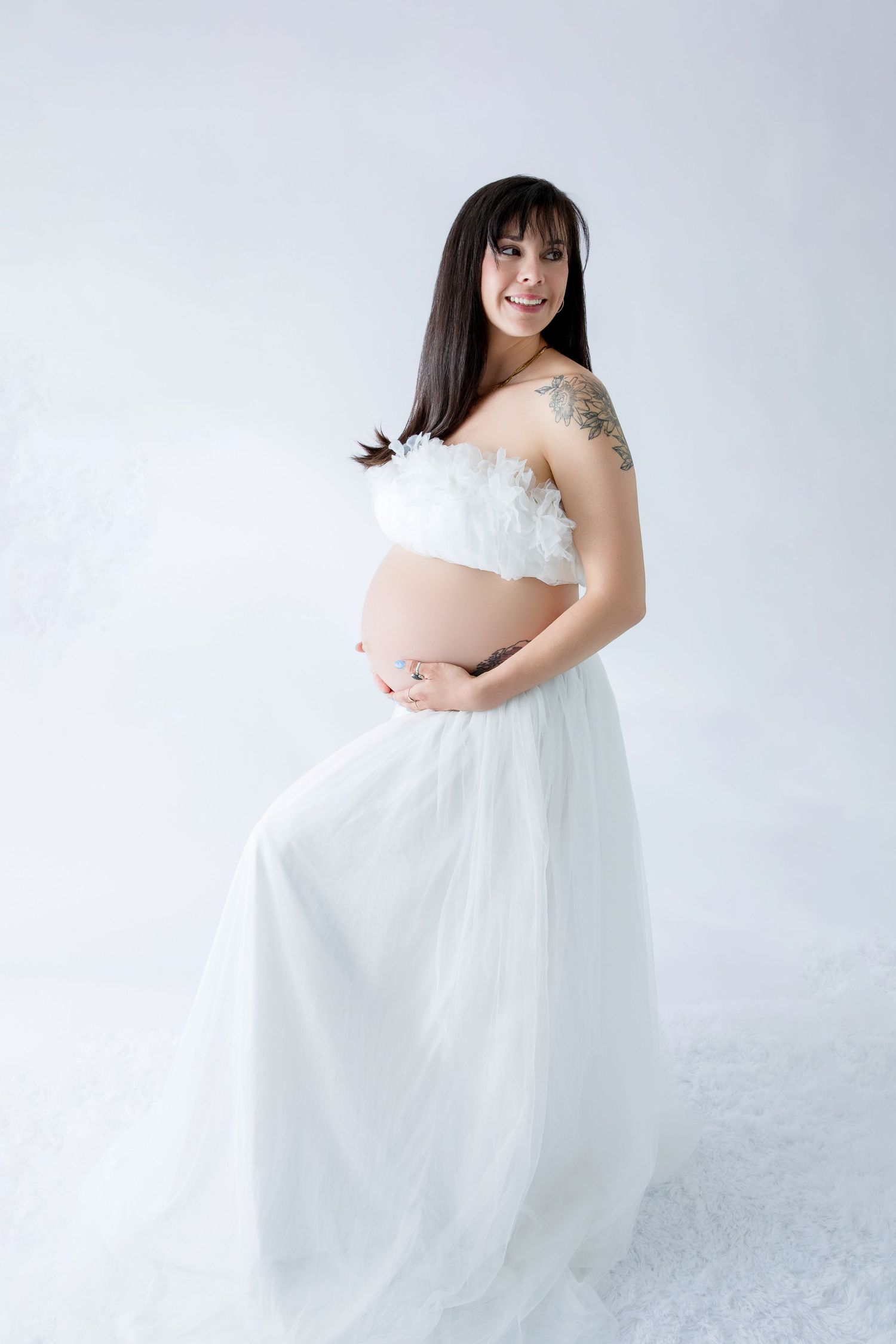 Smiling pregnant woman in white strapless gown with floral top cradling baby bump against white background.
