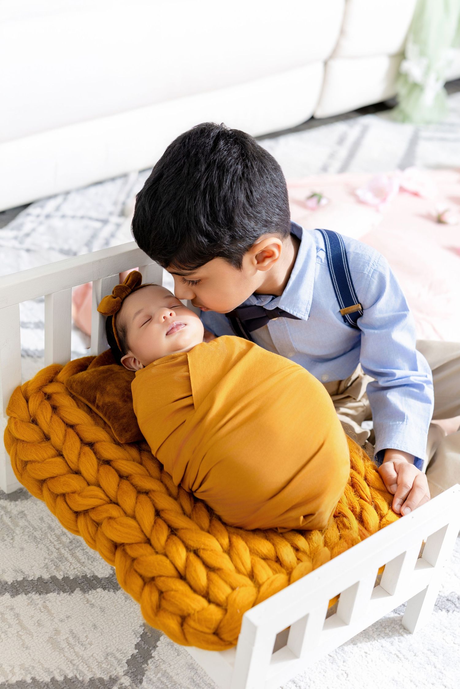 An older boy tenderly kisses a swaddled newborn baby lying on a chunky knit blanket in a small white crib.