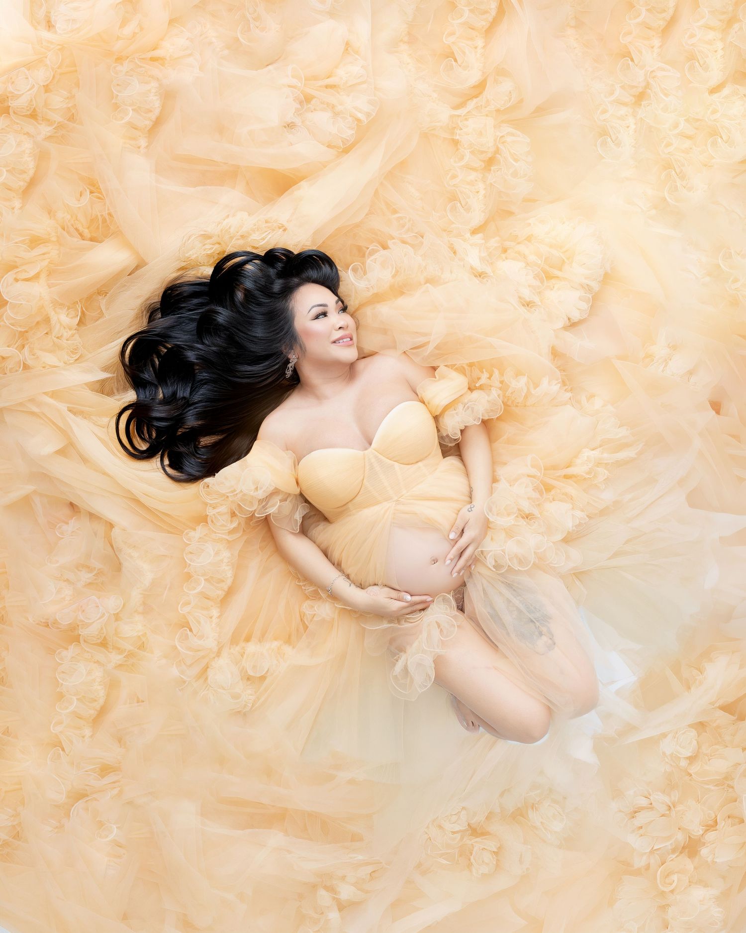 Pregnant woman in yellow bustier lying on flowy golden fabric, cradling baby bump, aerial maternity photography.