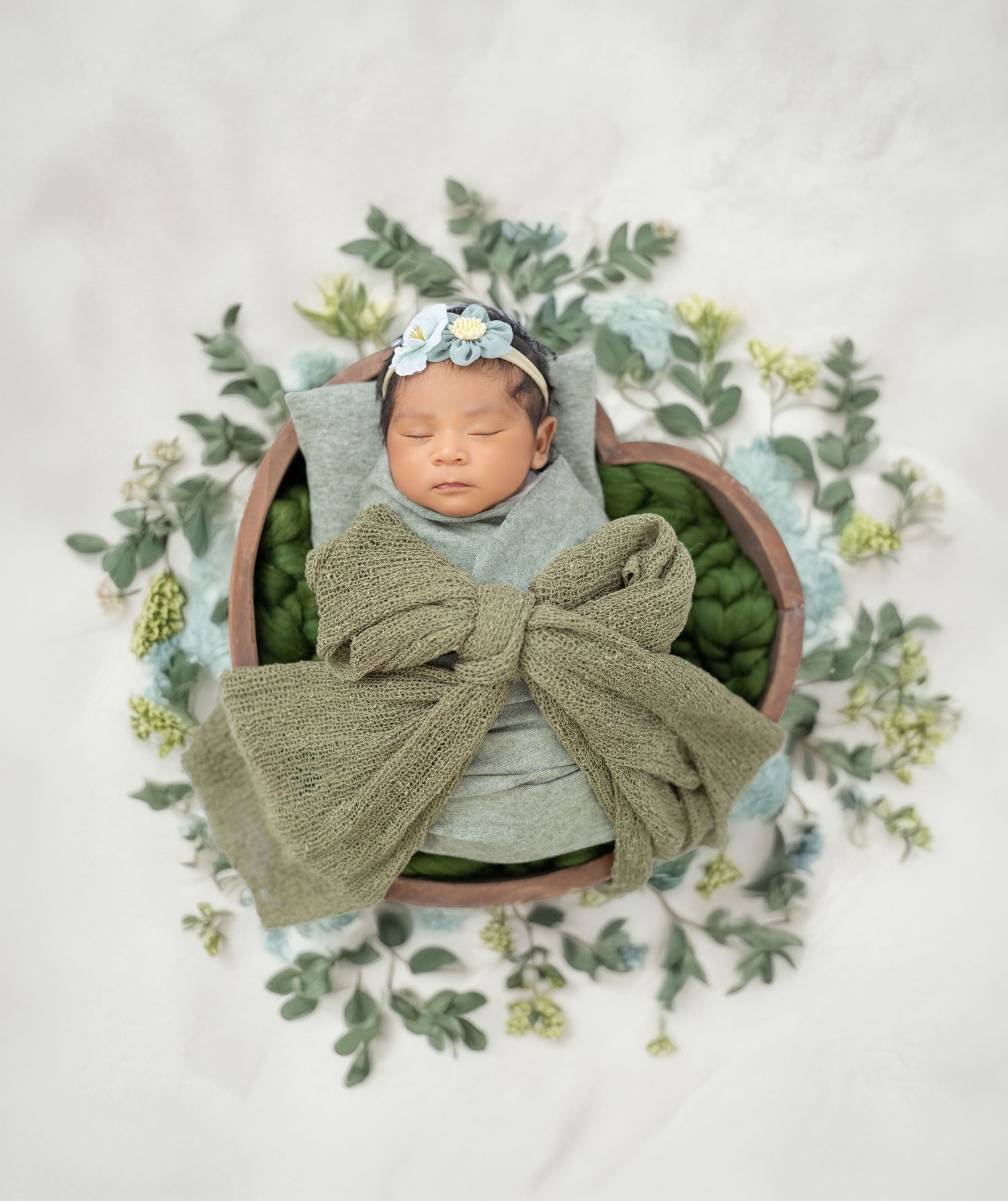 Sleeping newborn in sage green swaddle with bow resting in wooden bowl surrounded by eucalyptus leaves.