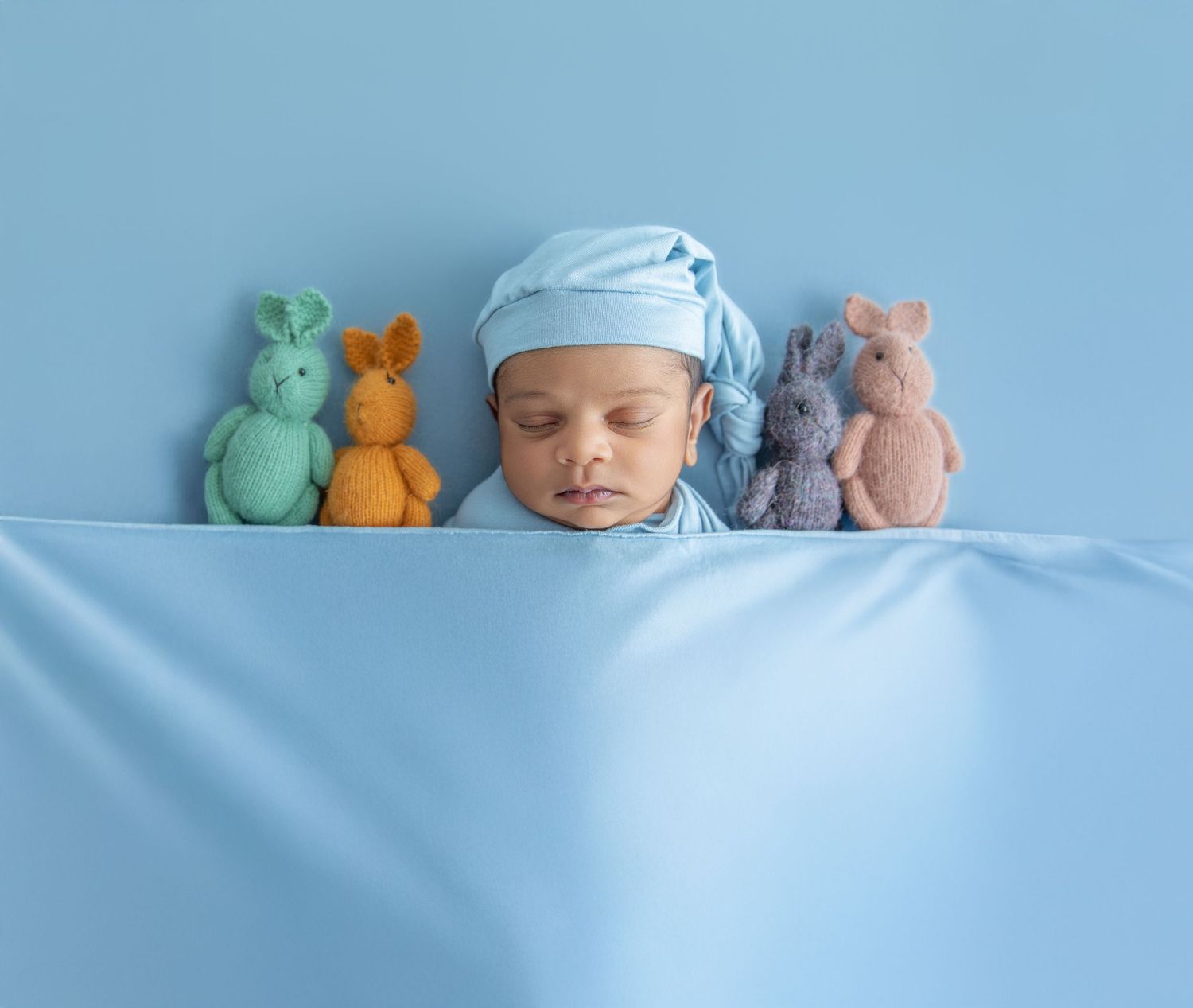 A sleeping baby in blue cap rests alongside colorful stuffed animal toys on blue bedding.