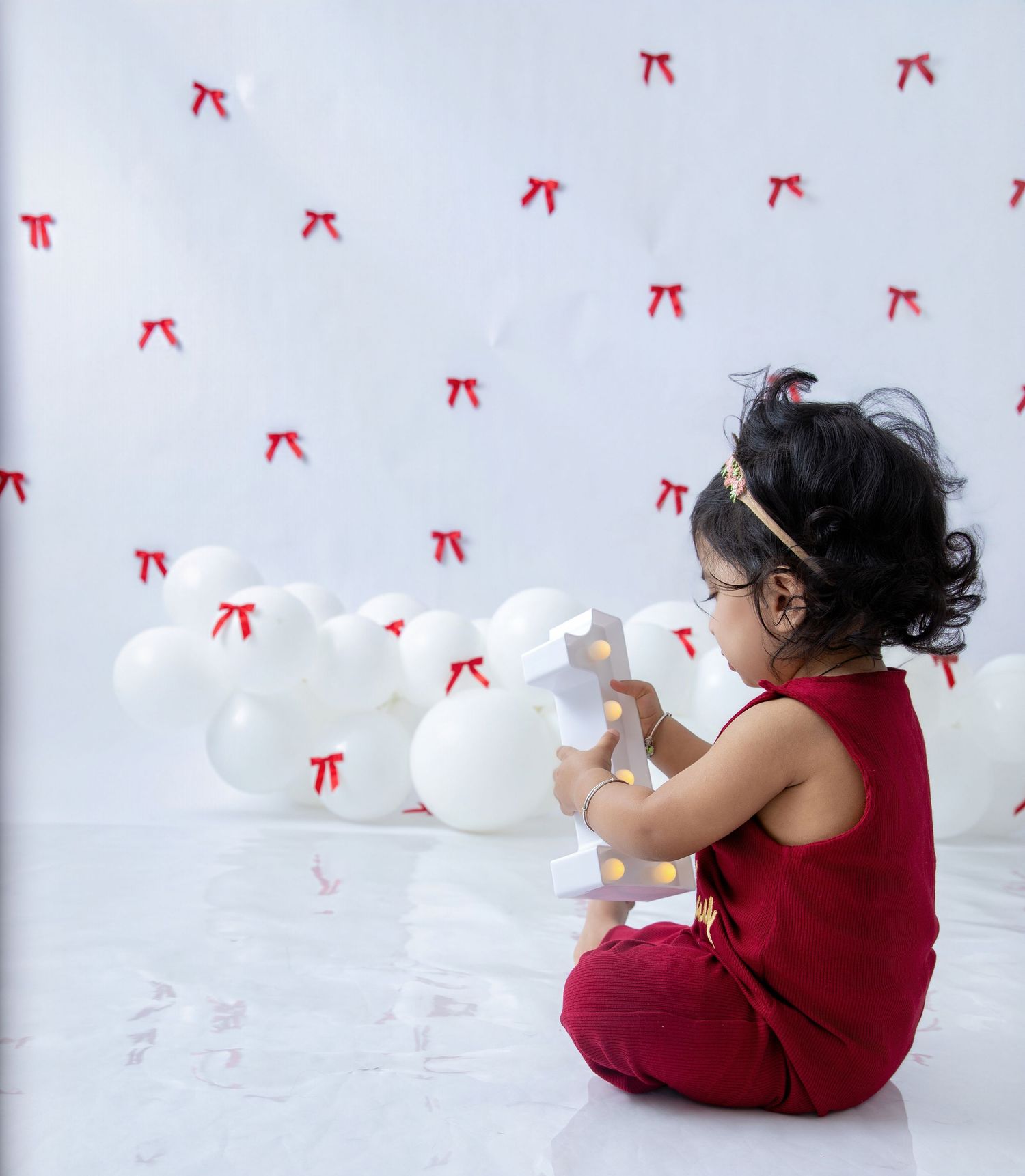 Baby in red romper sits among white balloons against decorated backdrop.