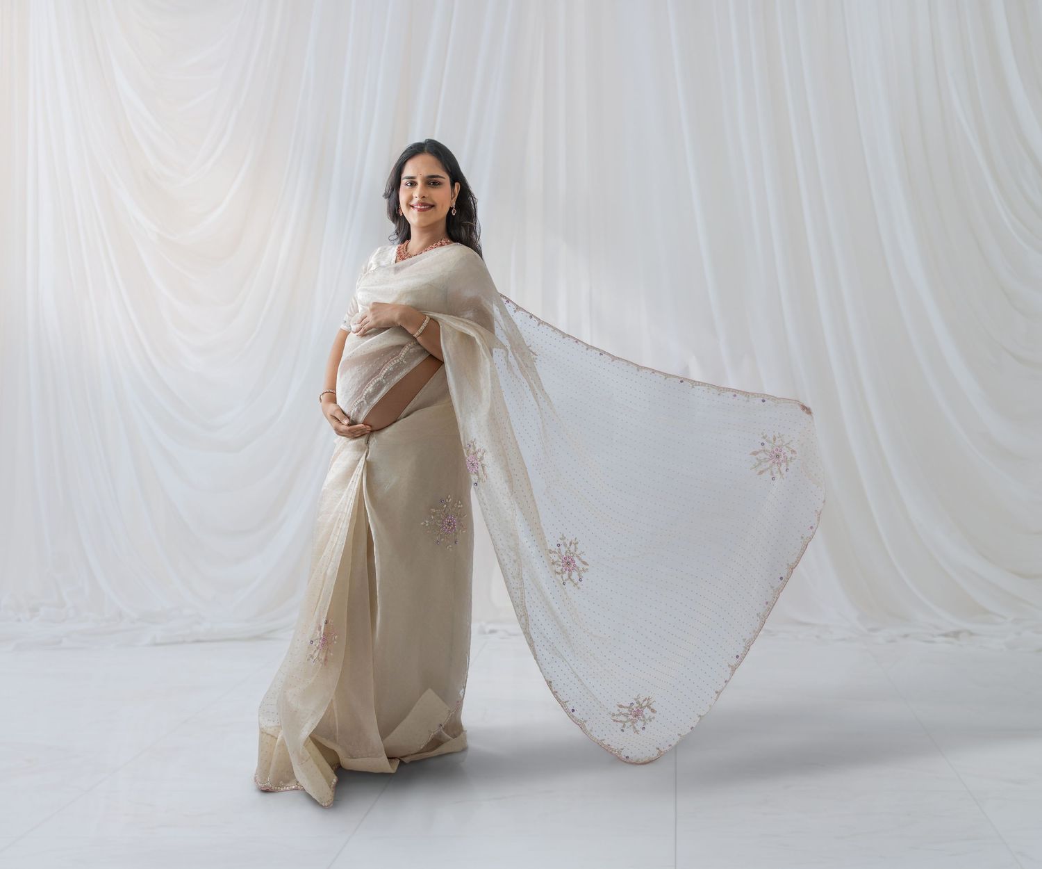 Pregnant woman in elegant white embroidered saree posing gracefully against a white draped backdrop.