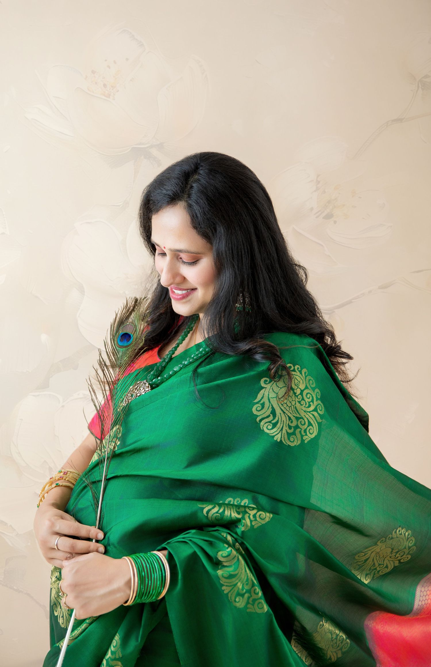 Pregnant woman in green silk saree holding peacock feather against floral wall.