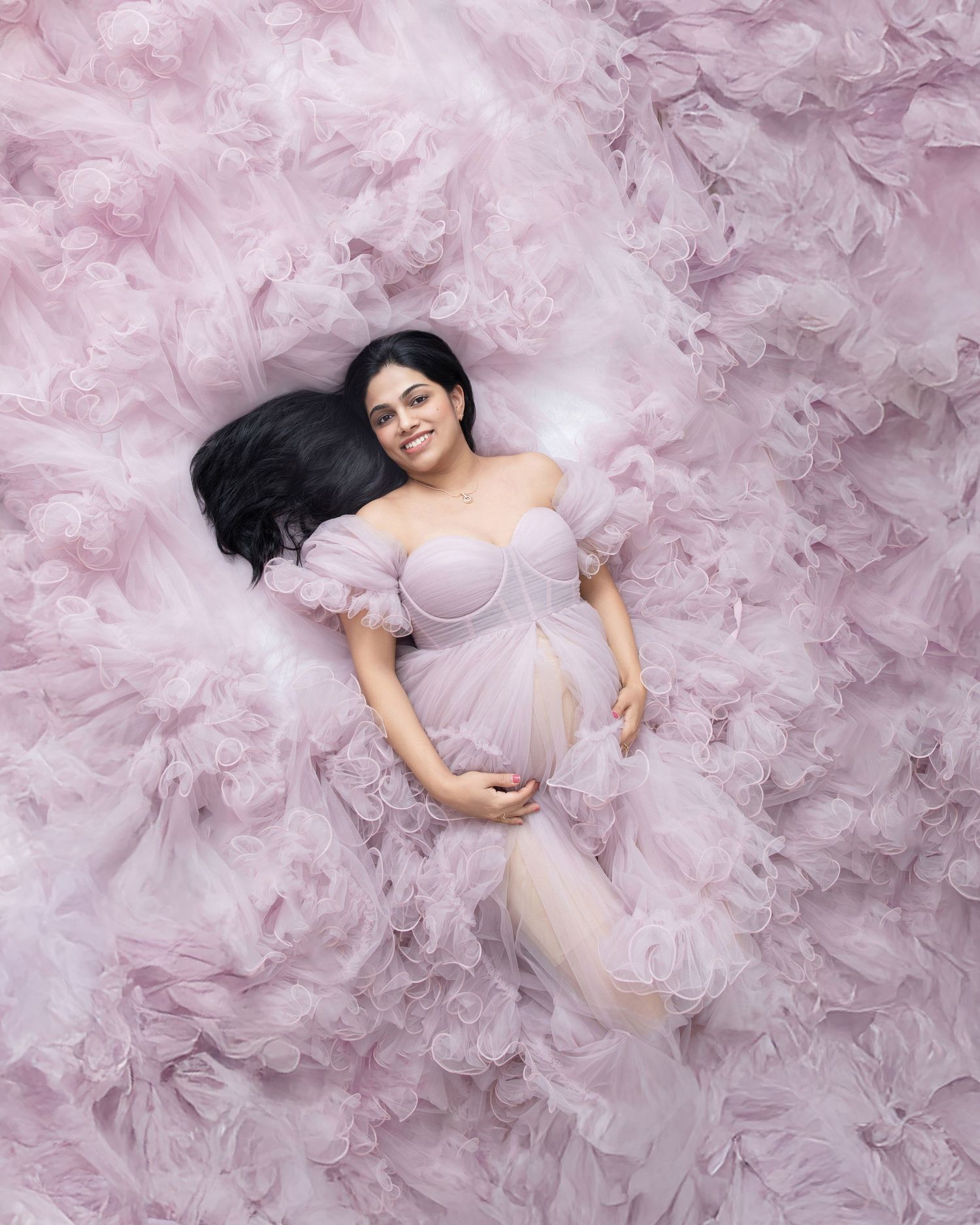 Pregnant woman in pink tulle gown lying gracefully surrounded by ruffled fabric in a dreamy maternity photo shoot.