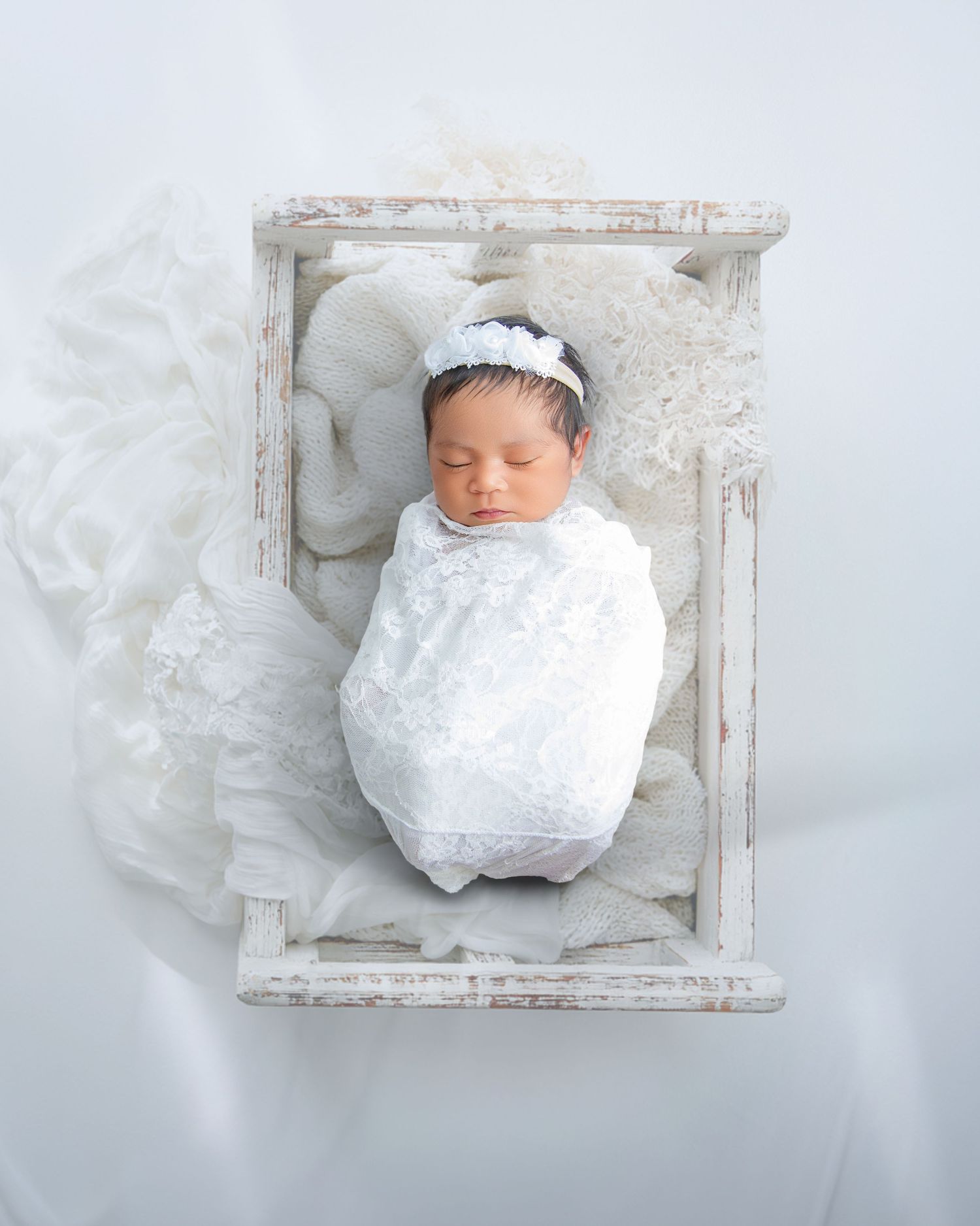 Newborn baby swaddled in white lace wrap with floral headband, sleeping peacefully in a rustic white wooden crate.
