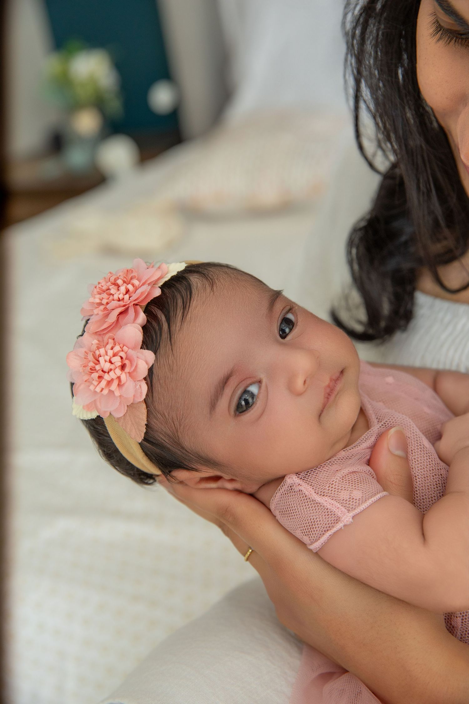 Baby wearing pink outfit with coral flower headband lying on white bedding.