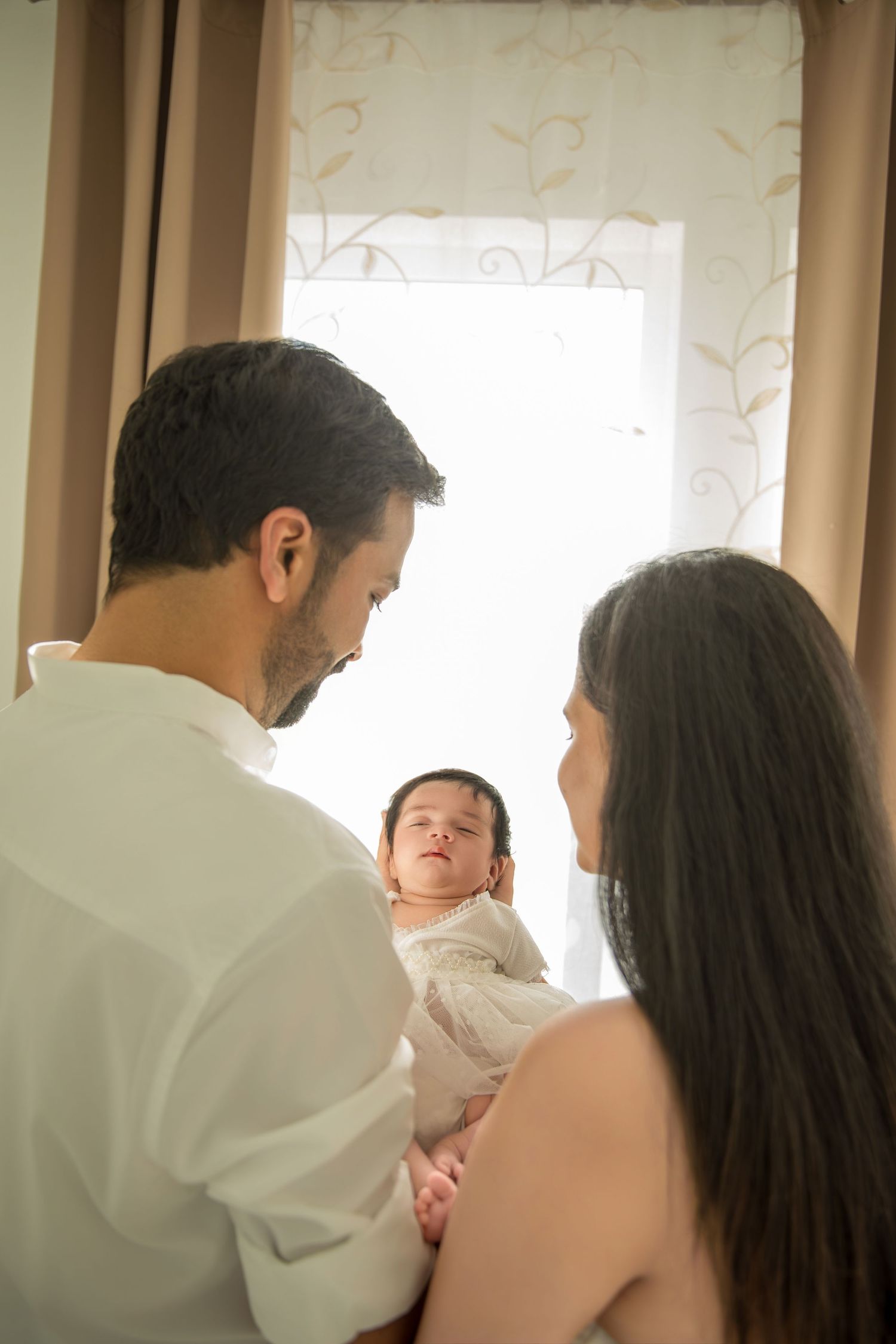 A tender family moment as parents cradle their sleeping newborn baby against a soft window-lit background.