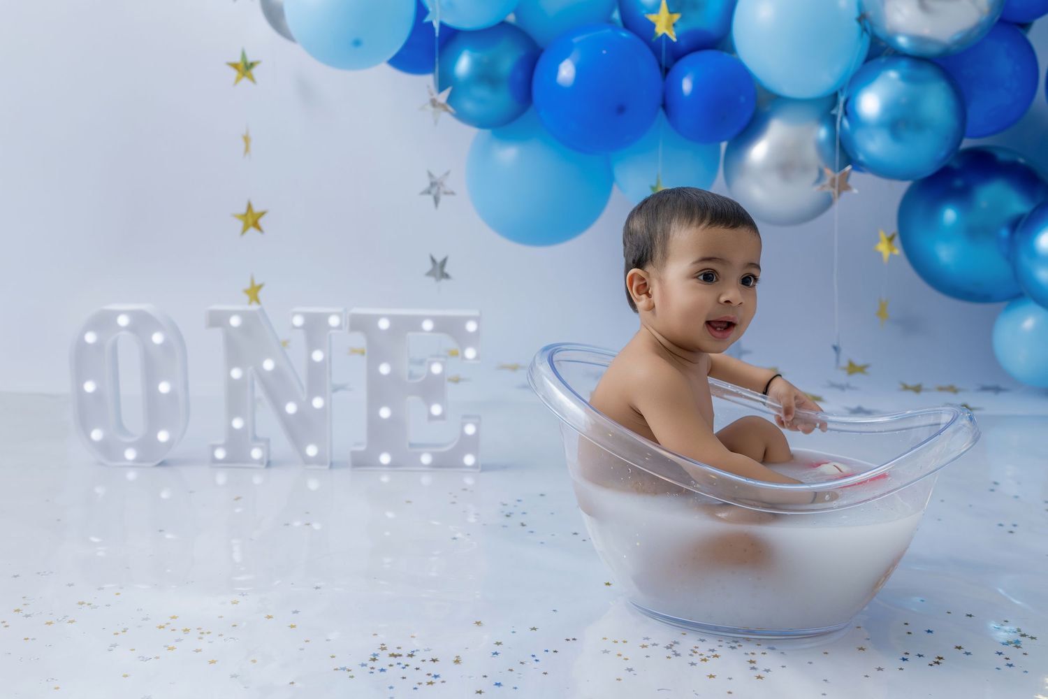 A milk bath photoshoot setup with blue balloon arch and glowing ONE sign against white backdrop.