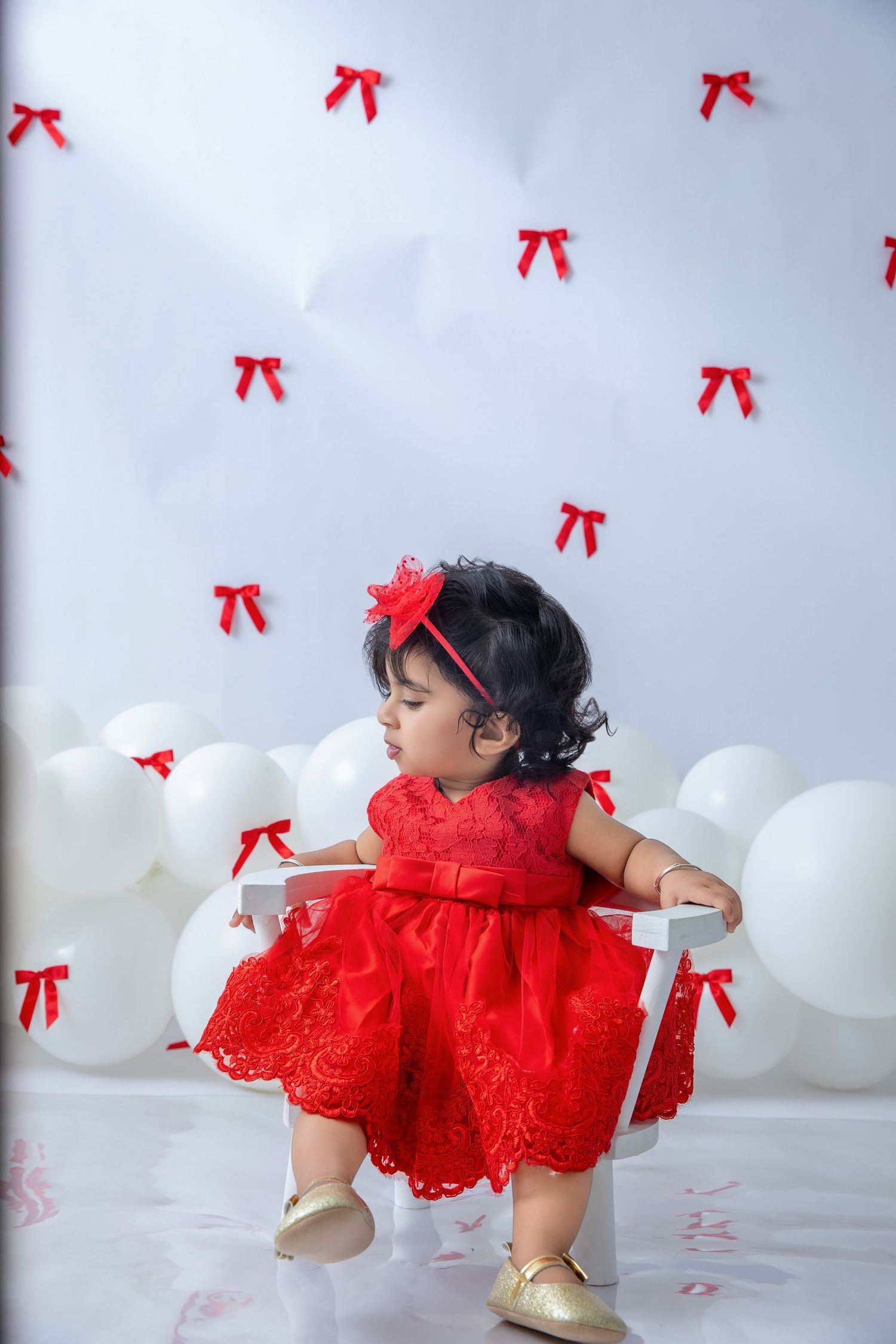 Toddler in red lace dress twirls against white backdrop with red bows and balloons.