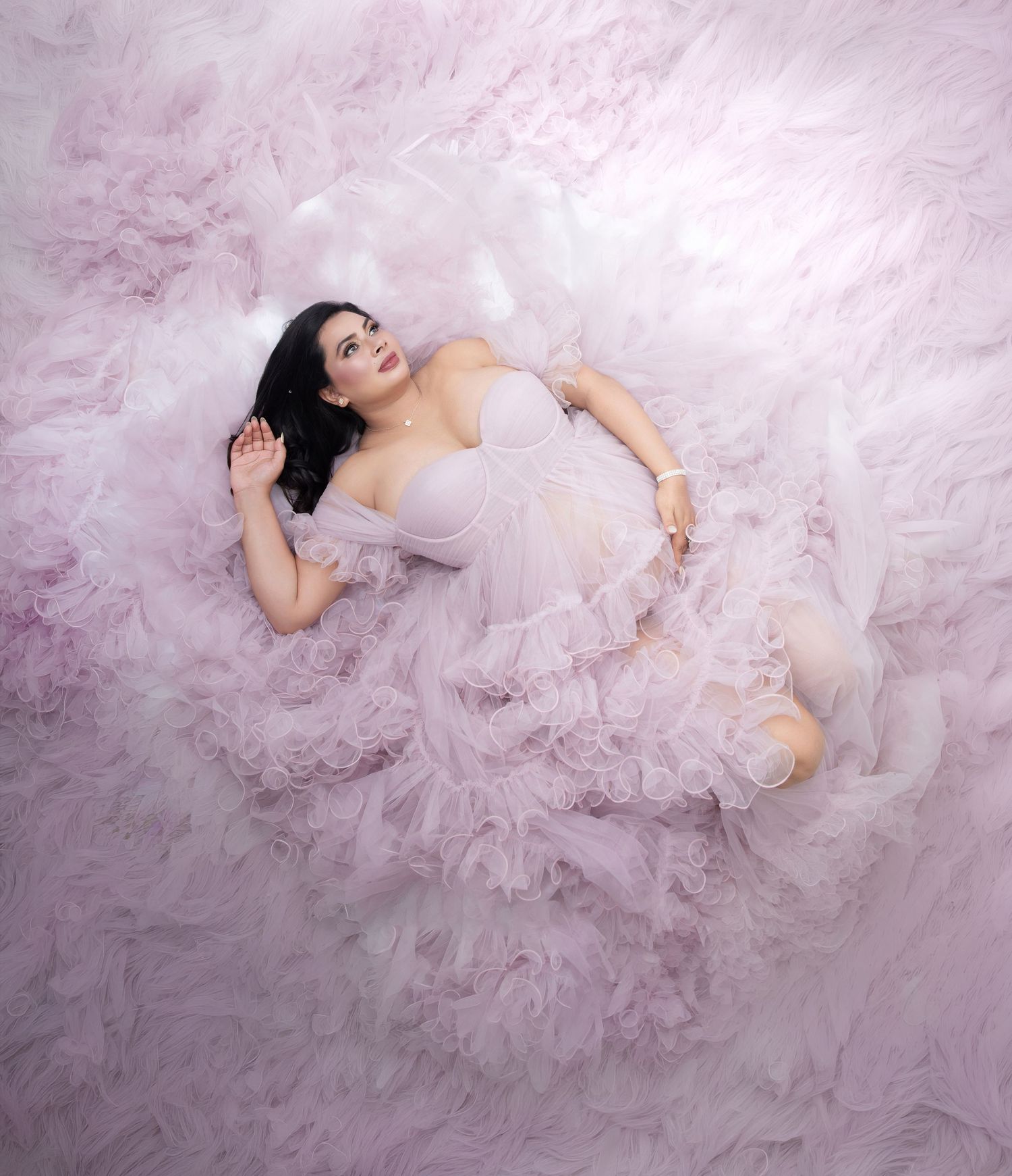 Woman in pink tulle gown lying gracefully on a soft pink ruffled backdrop in an ethereal fashion portrait.