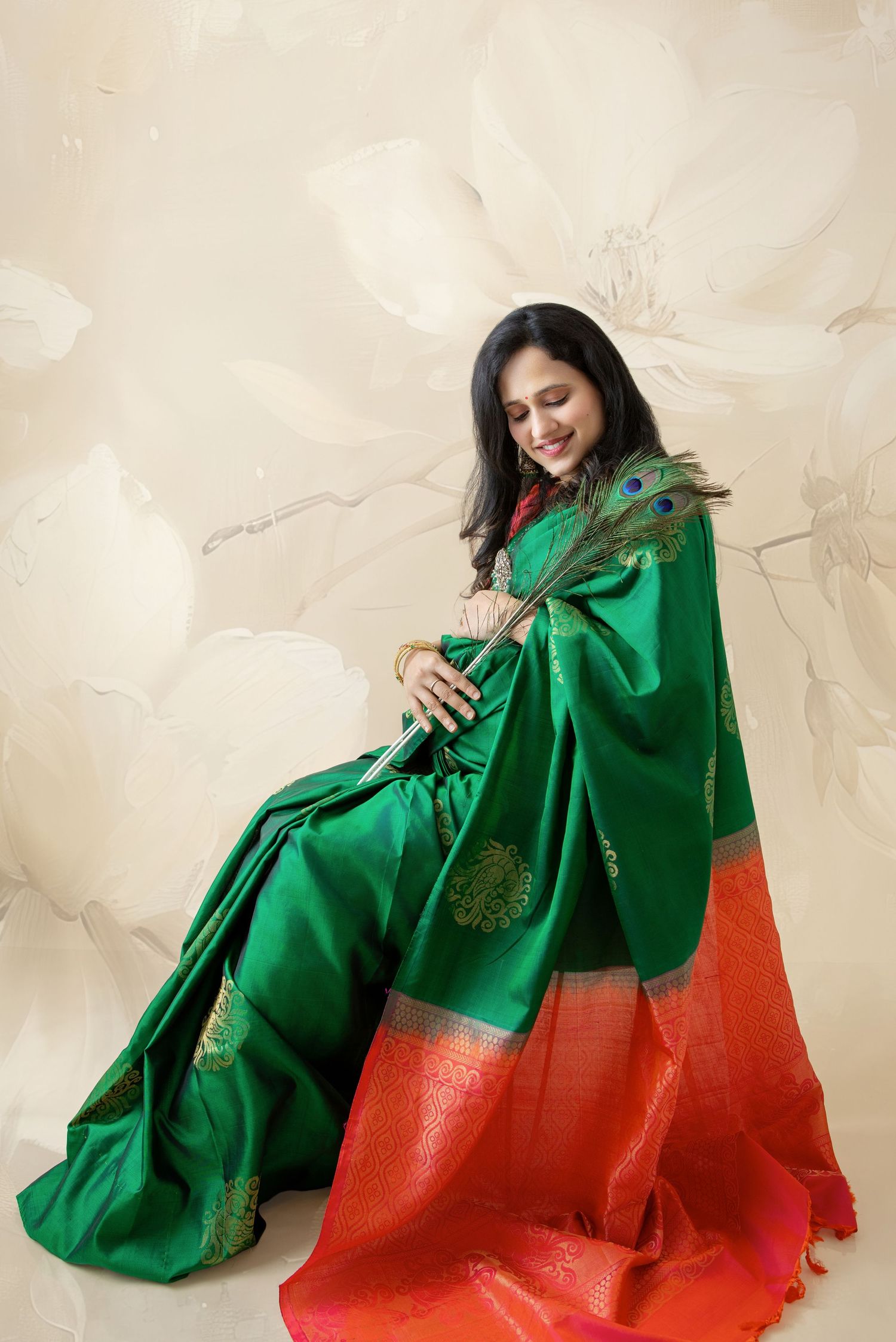 Indian woman in elegant green and red silk saree holding peacock feather during traditional photoshoot.