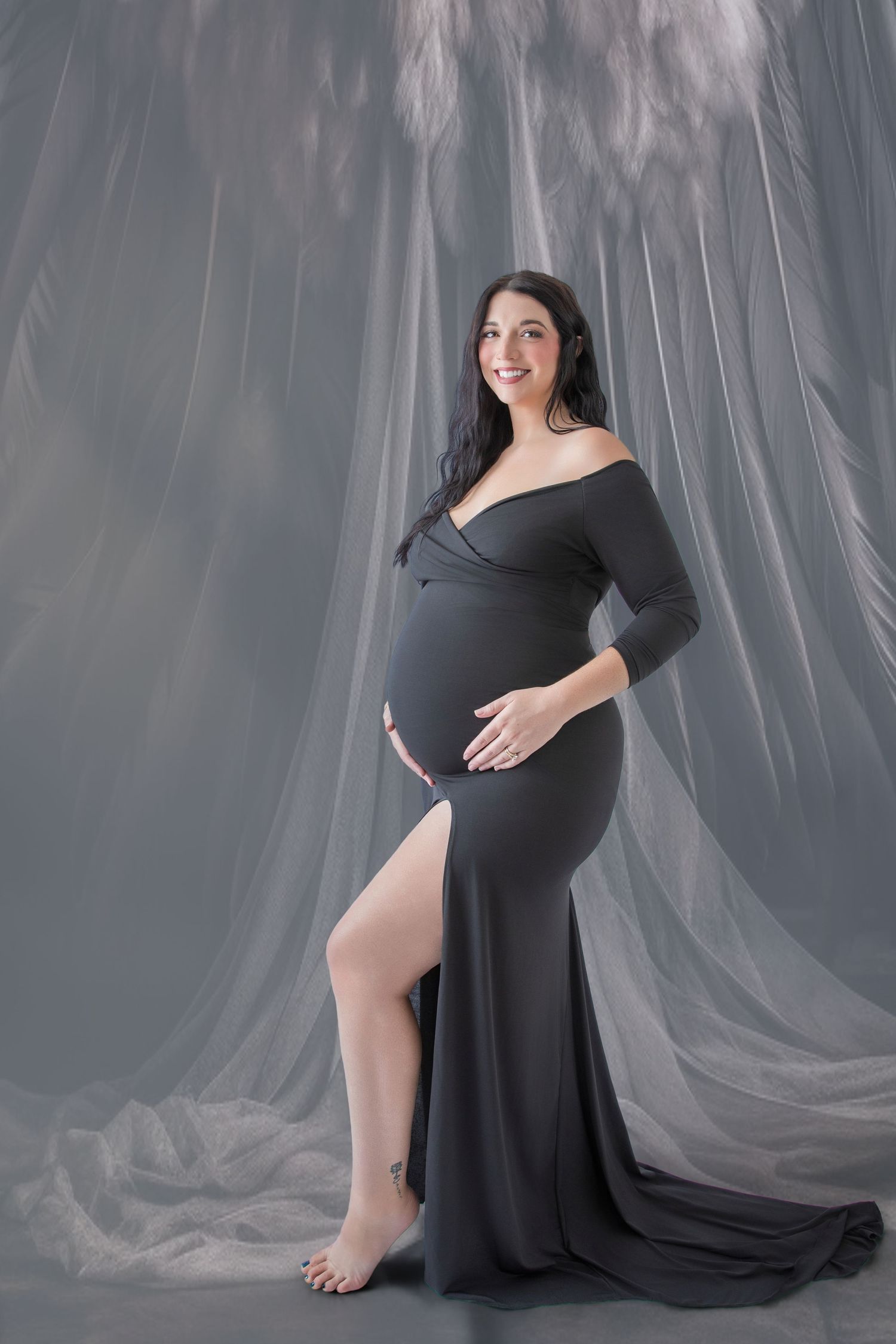 Smiling pregnant woman in elegant off-shoulder gray maternity gown posing in studio with flowing sheer backdrop.