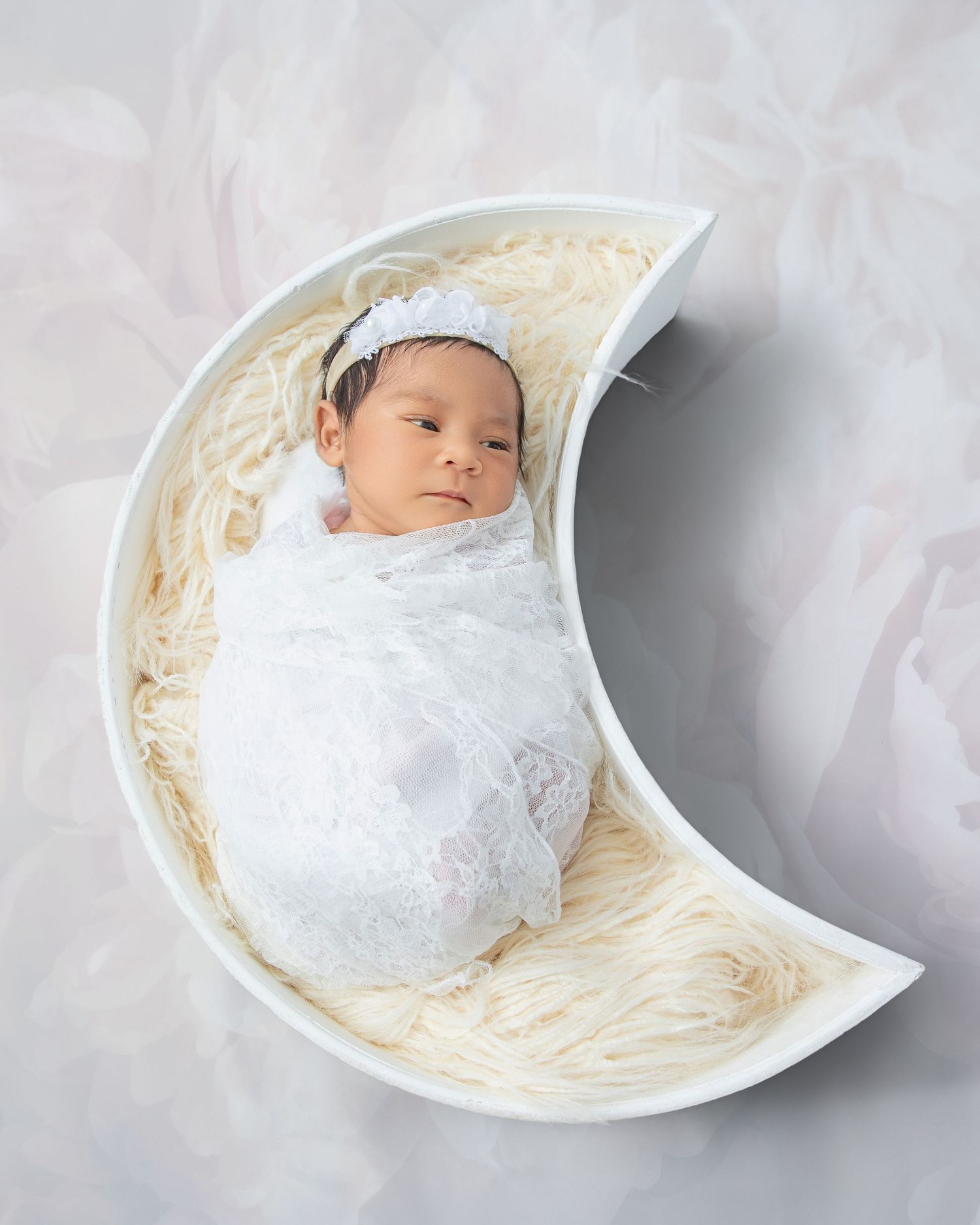 Newborn baby swaddled in white lace wrap resting in a crescent moon prop with cream fur filler.