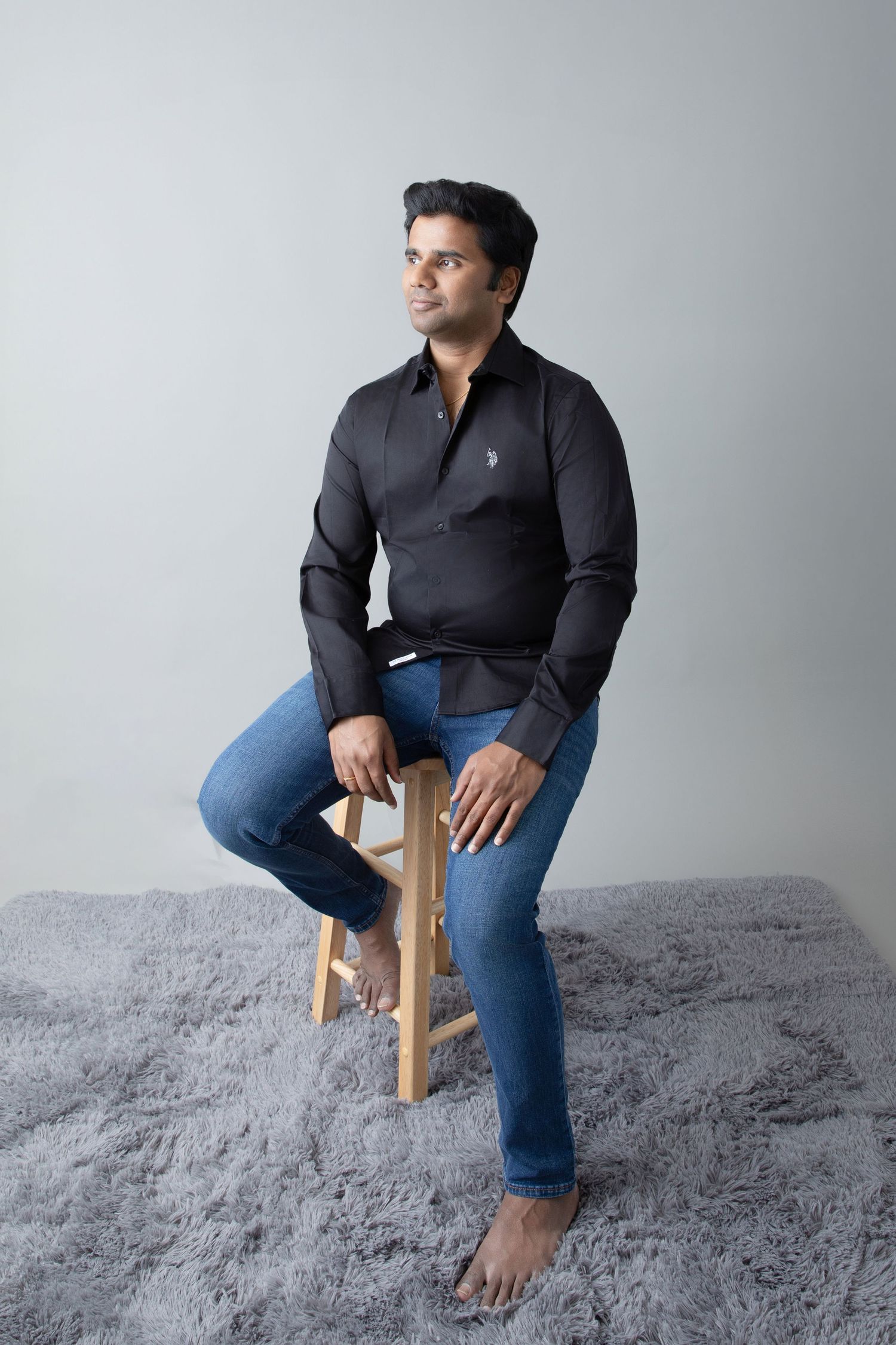 Man in dark polo shirt and jeans sitting casually on wooden stool against gray background.