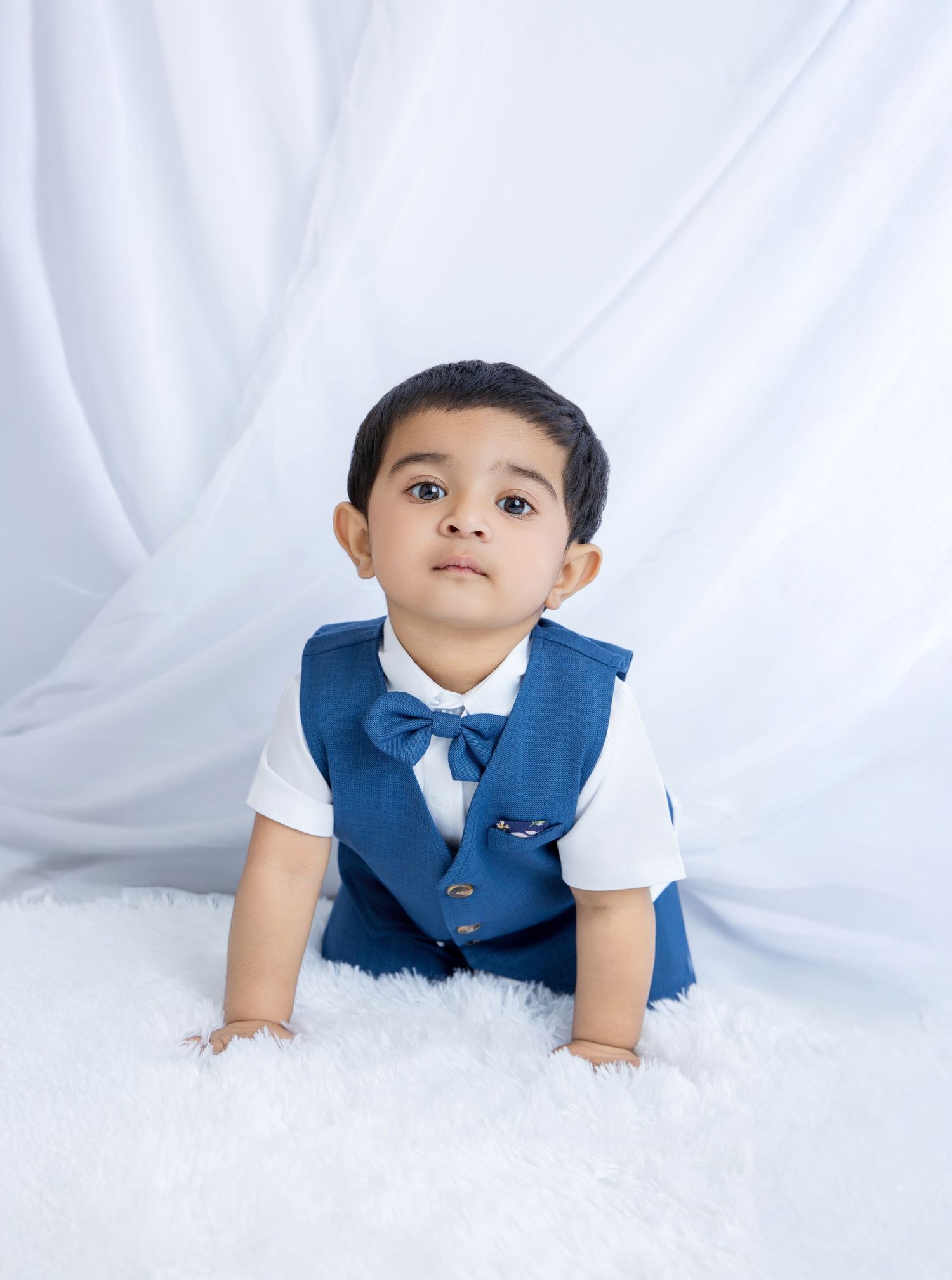 A small child in a blue vest and white shirt sits on a white fluffy carpet against a light backdrop.