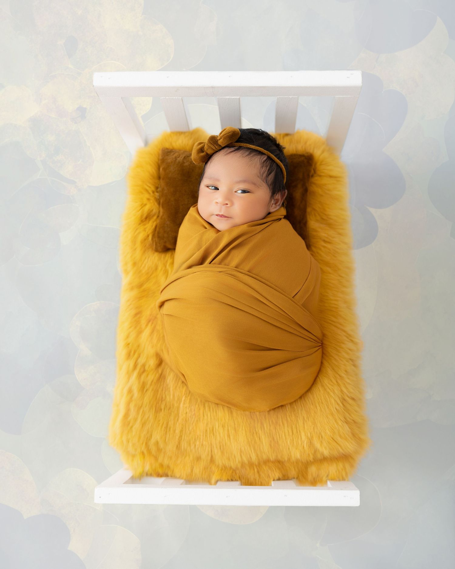 Newborn baby swaddled in mustard yellow wrap, resting on fluffy fur in a tiny white crib for a studio photo.