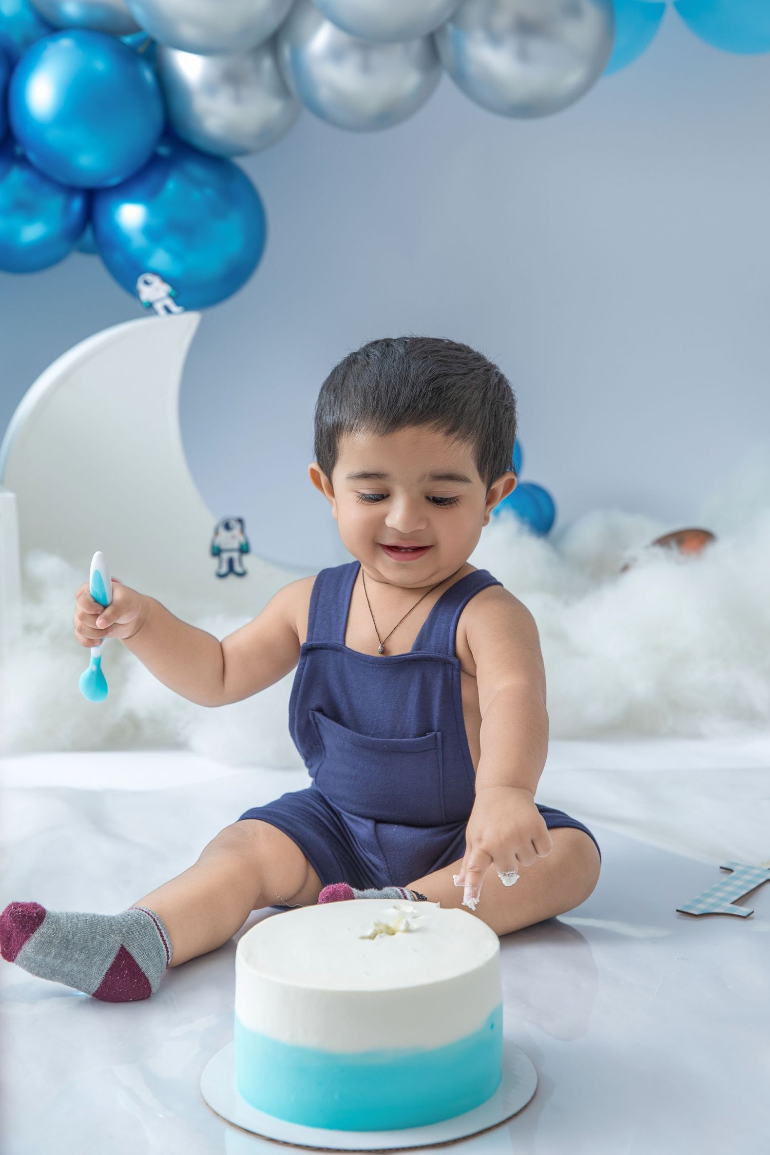 Small child in navy outfit reaches for white and turquoise cake during photo session with balloon backdrop.