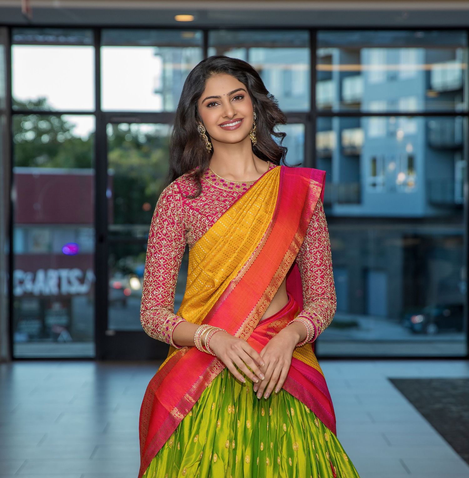 Person wearing vibrant traditional Indian attire with yellow and pink colors poses in front of modern building exterior.