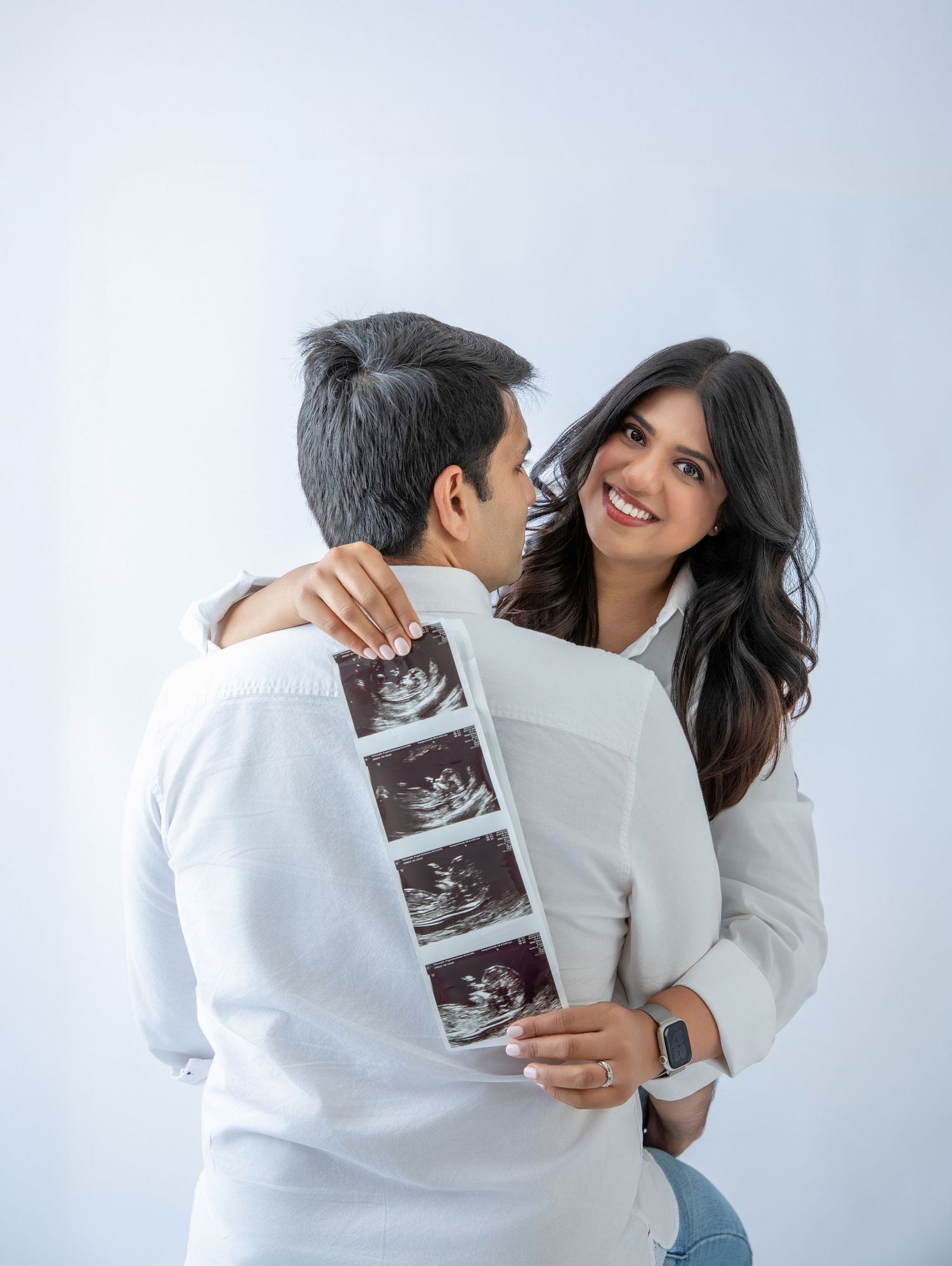 Happy couple embracing and holding ultrasound photos, announcing pregnancy in a bright white studio setting.