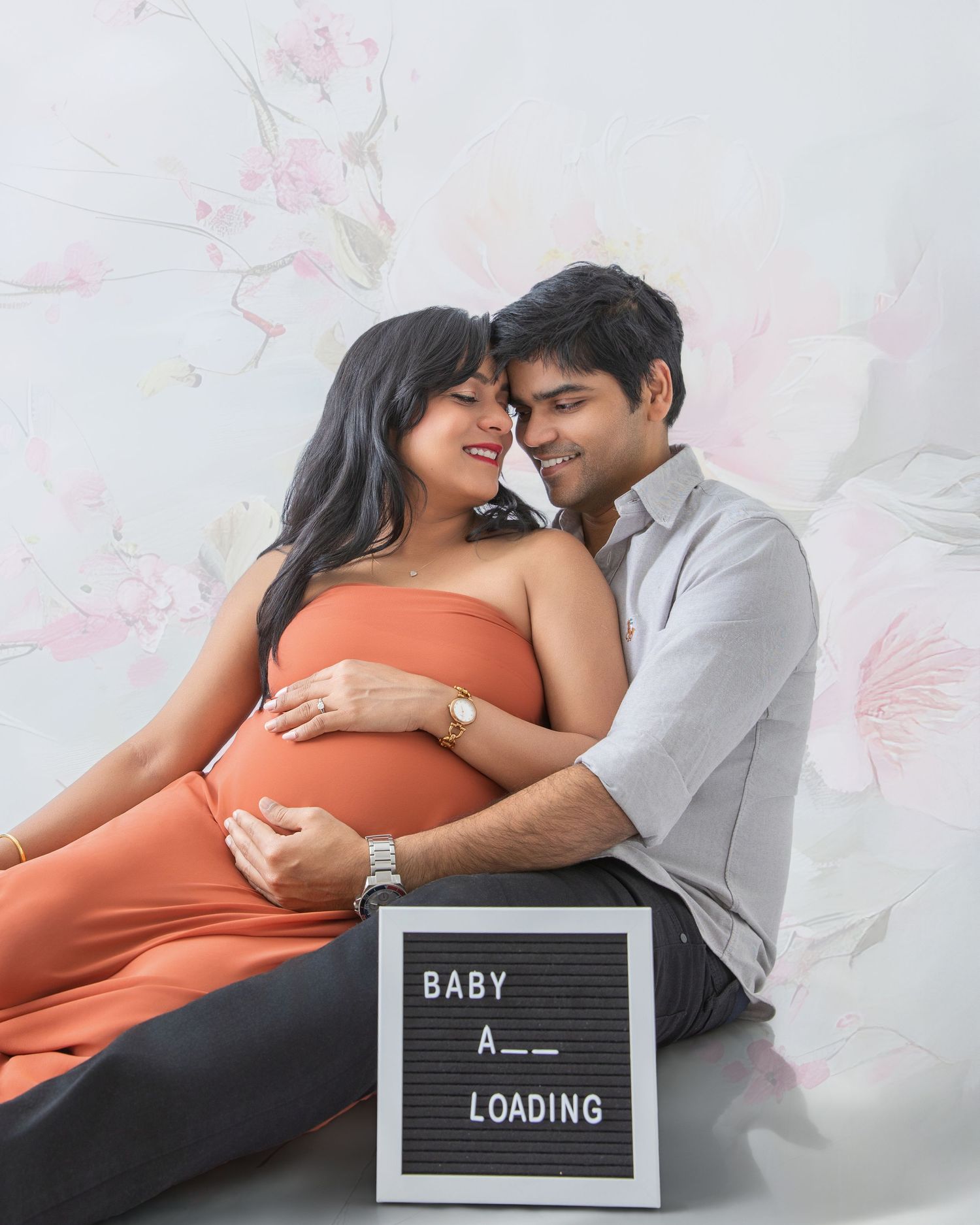 Pregnant couple posing for maternity photo shoot with Baby Loading sign, woman in orange dress, floral background.